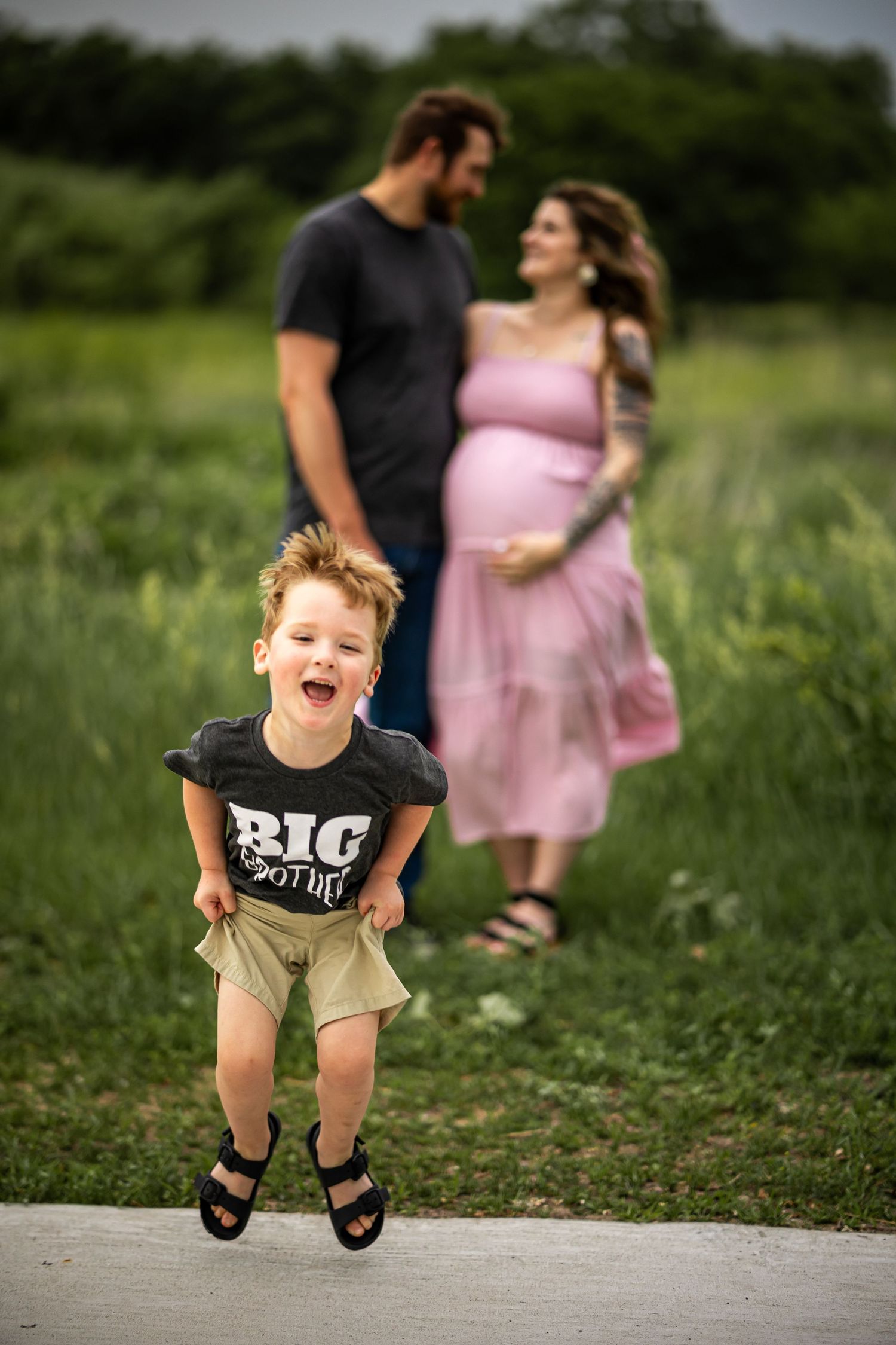 Excited boy in Big Brother shirt jumps as pregnant mom in pink dress and dad share tender moment outdoors.