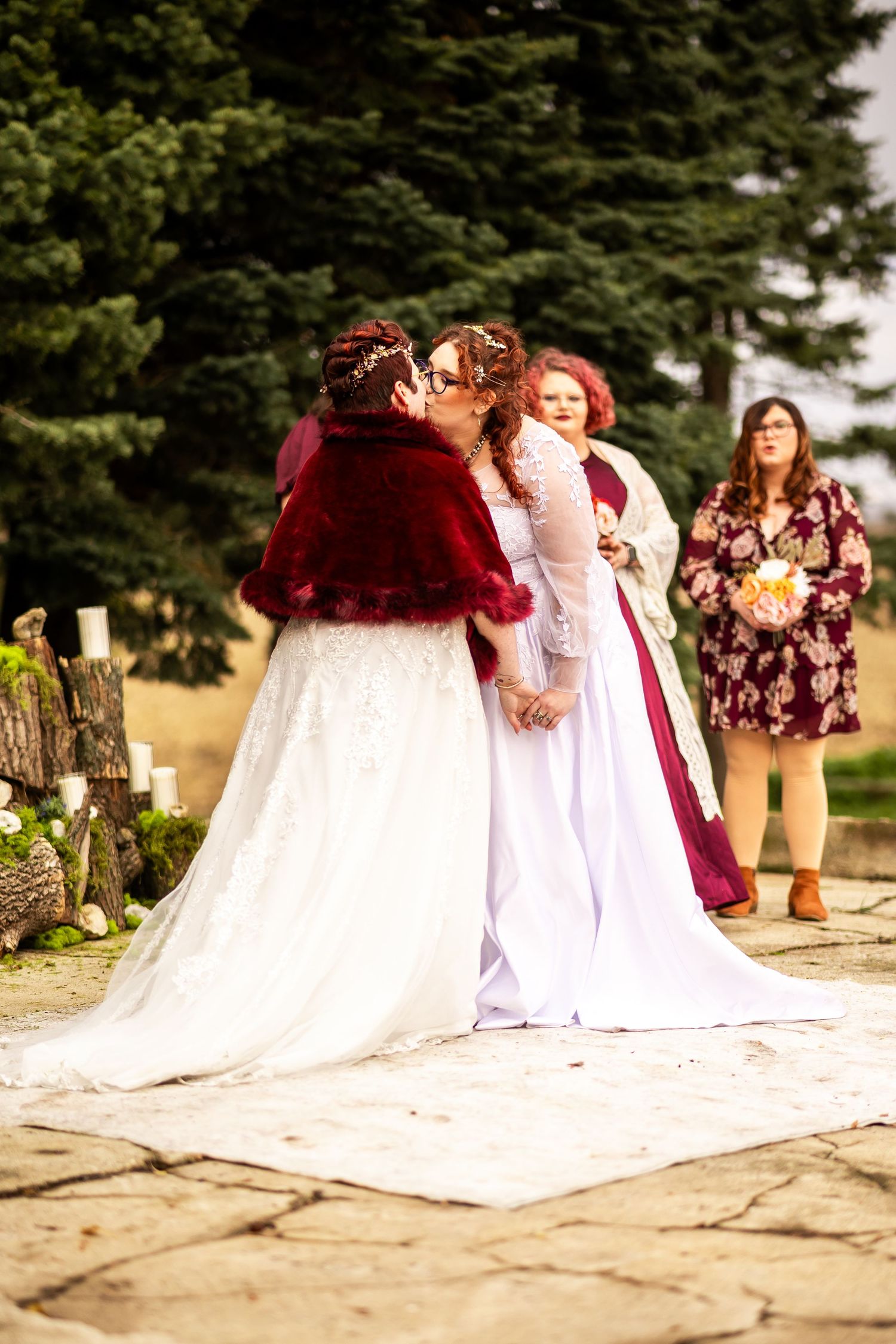 Two brides share a kiss at an outdoor wedding ceremony, one in white lace gown with red cape, guests watching behind.
