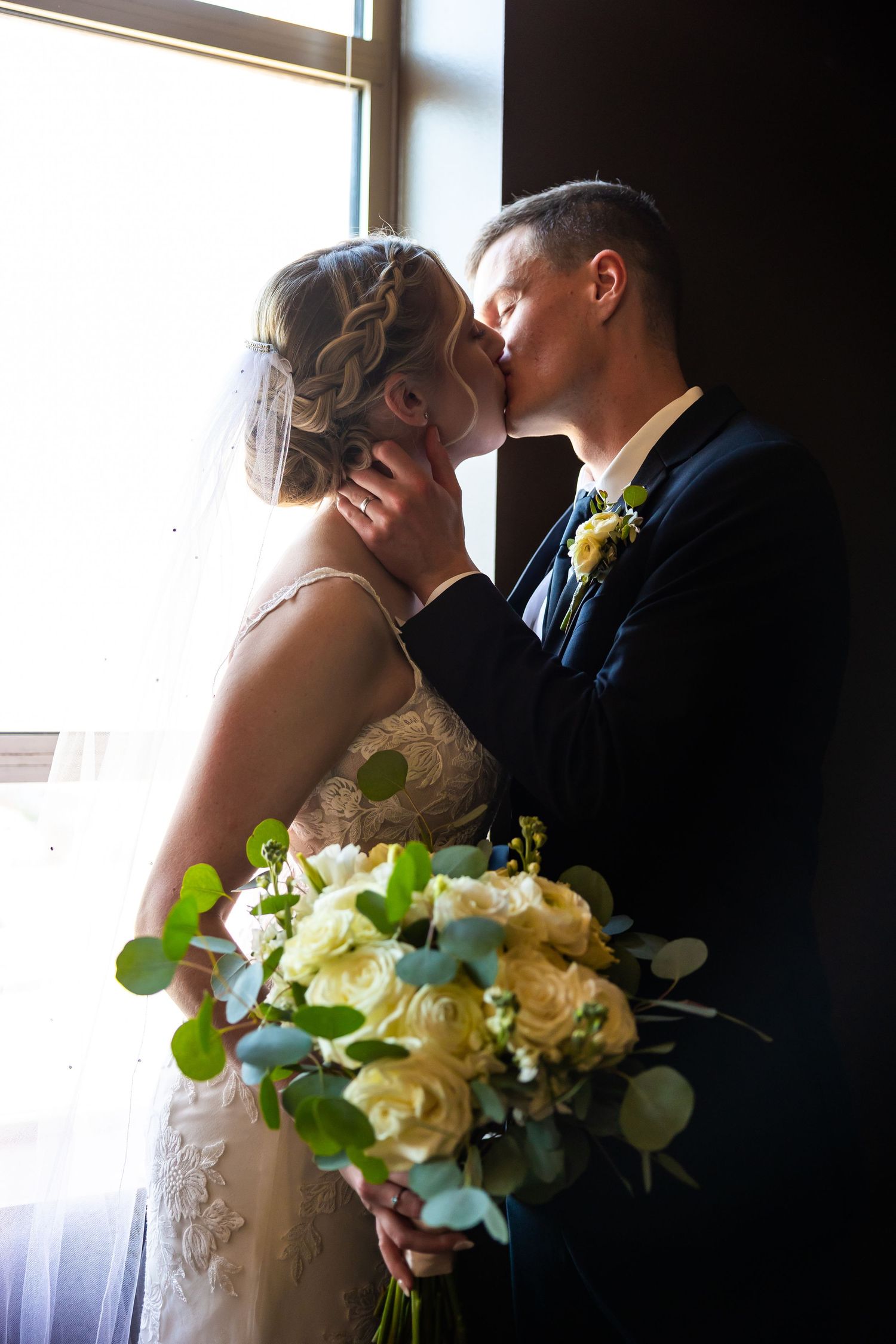 Newlyweds share romantic kiss by window with elegant white and green bouquet.