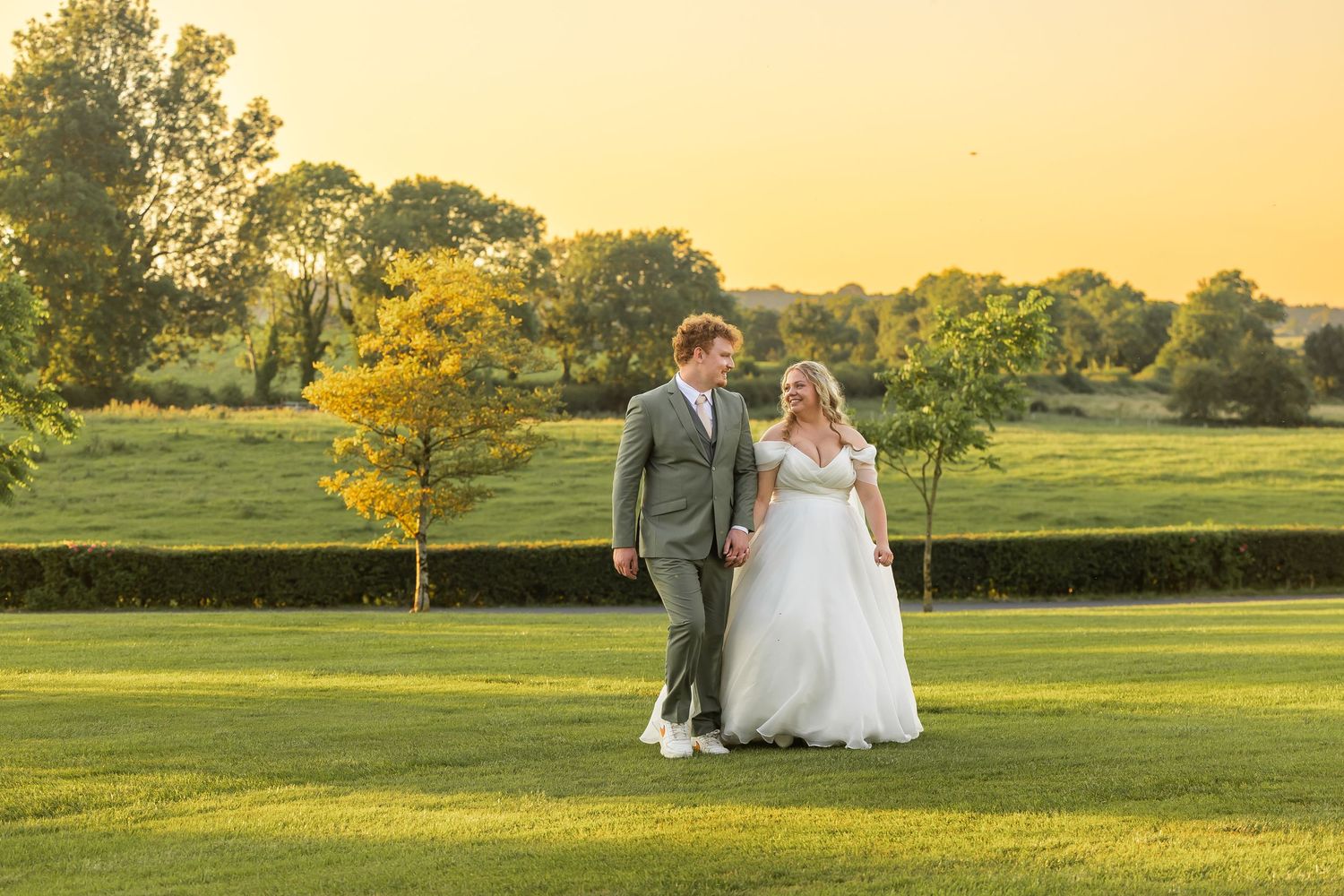A bride and groom walk hand in hand across lush green grounds at golden hour during their outdoor wedding celebration.