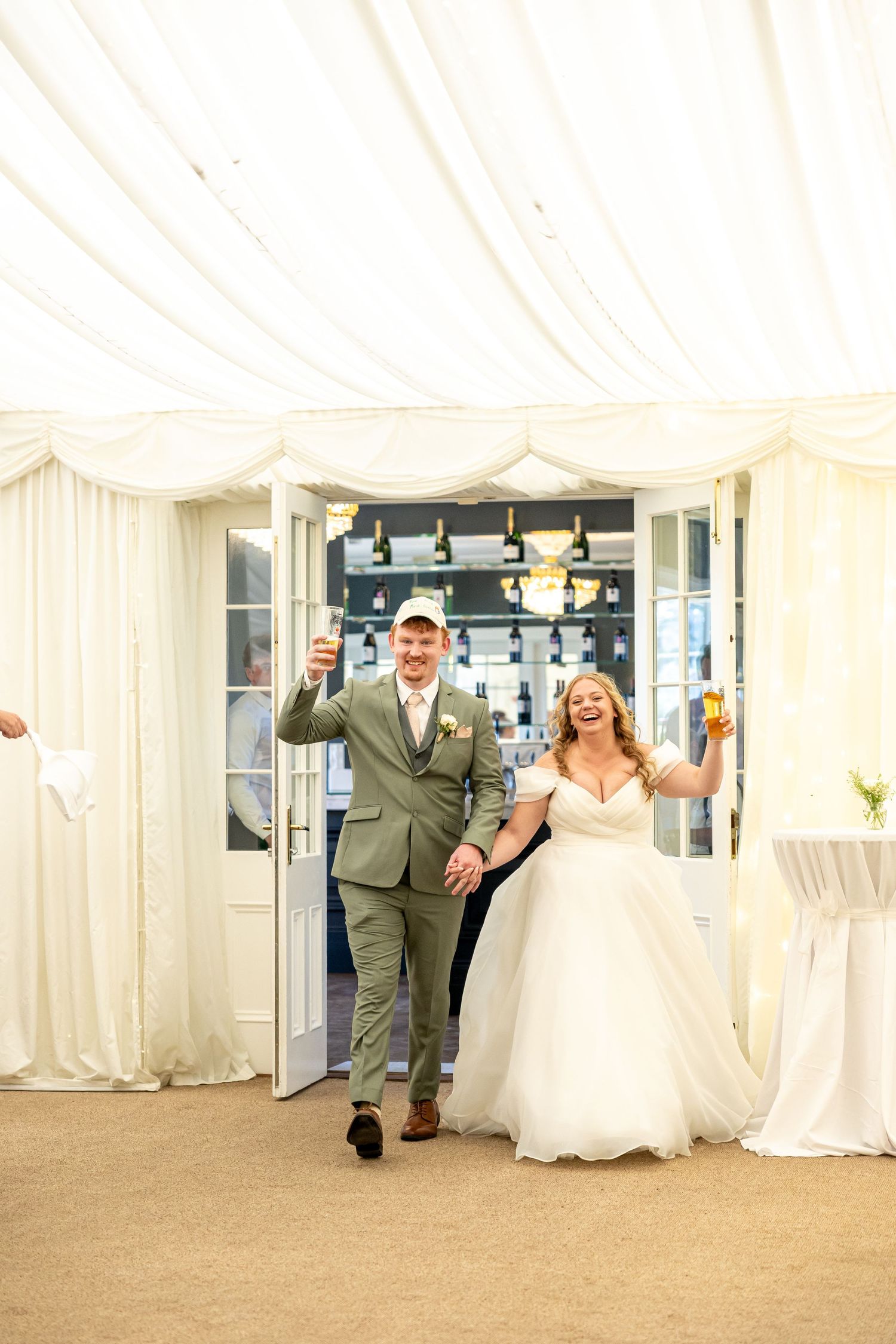 Bride and groom entering wedding reception marquee holding drinks, smiling joyfully in elegant attire.