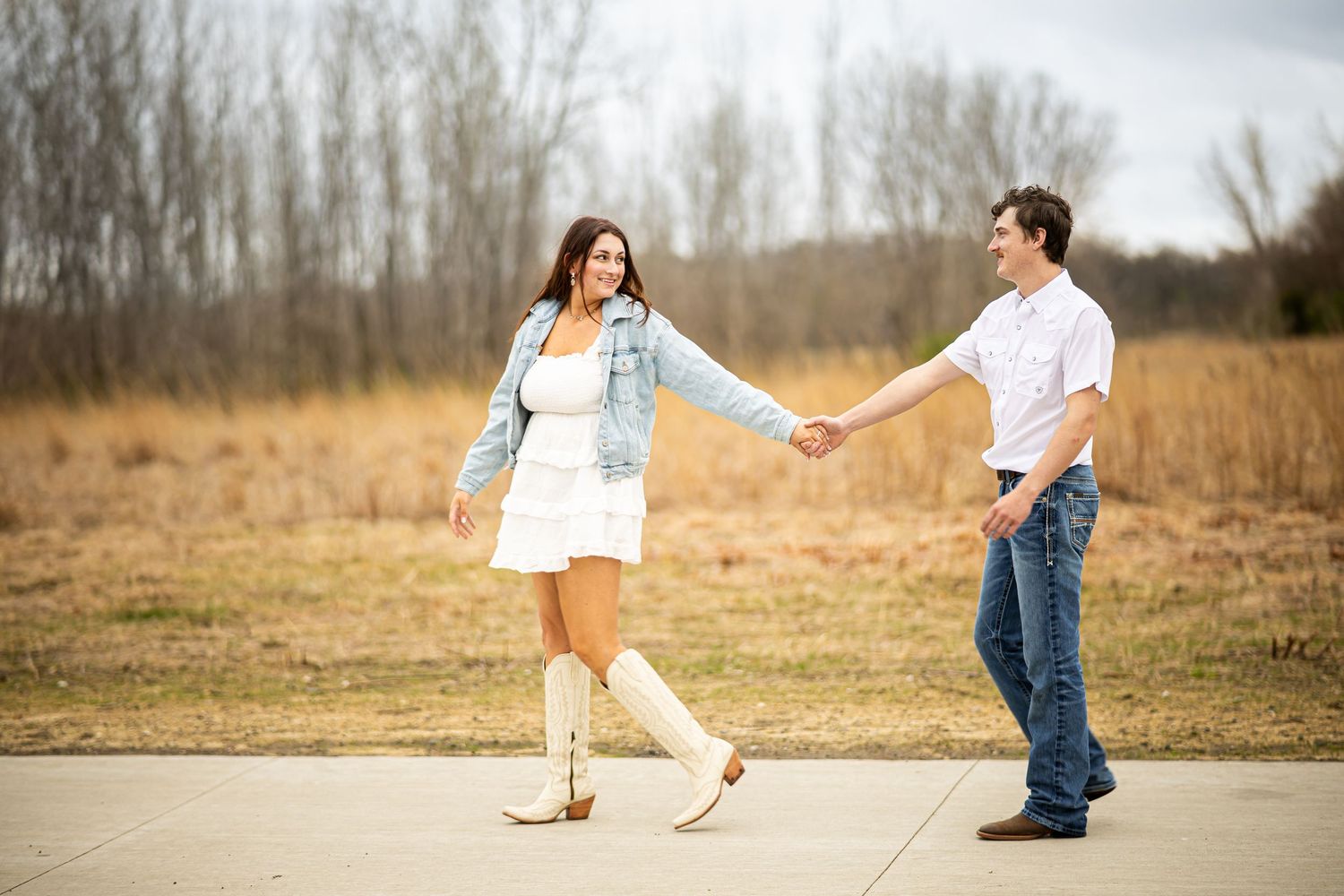 Couple holding hands and walking on a path in a field, woman in white dress and denim jacket with cowboy boots.