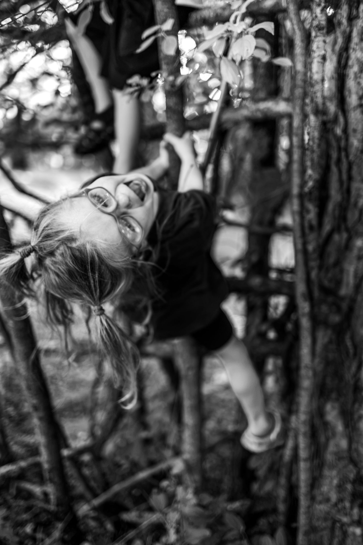 Black and white photo of a young girl with pigtails joyfully climbing a wooden ladder in a forest.