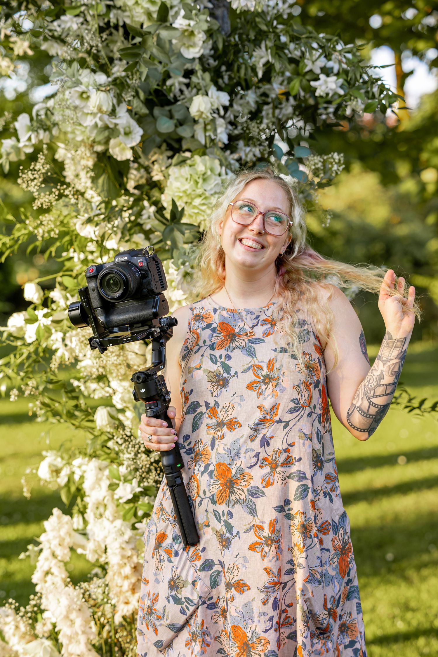 Smiling blonde woman with glasses holds camera gimbal outdoors near white floral arch, wearing floral dress with arm tattoo.