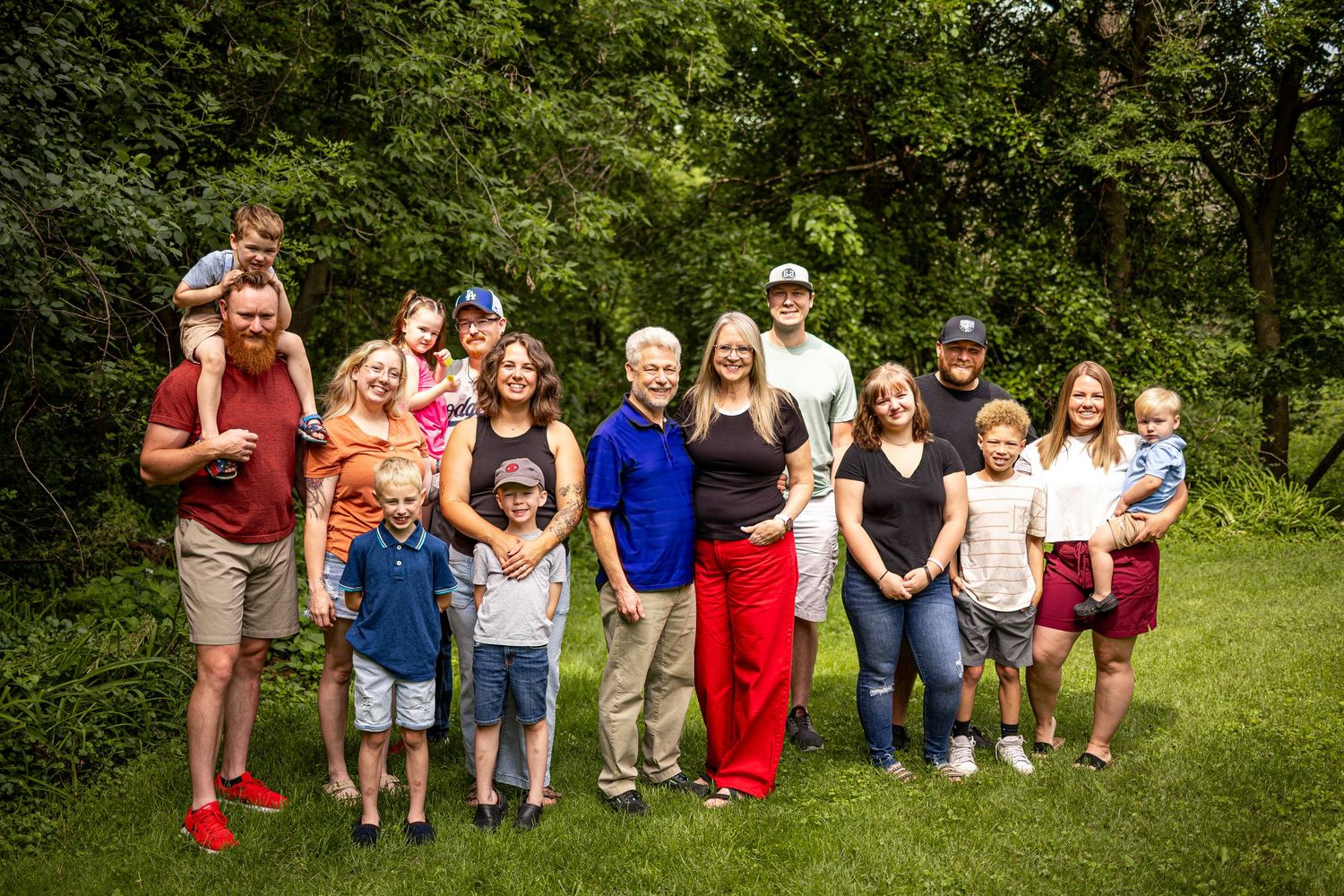 Large multigenerational family group smiling together outdoors on green grass with lush trees in background.