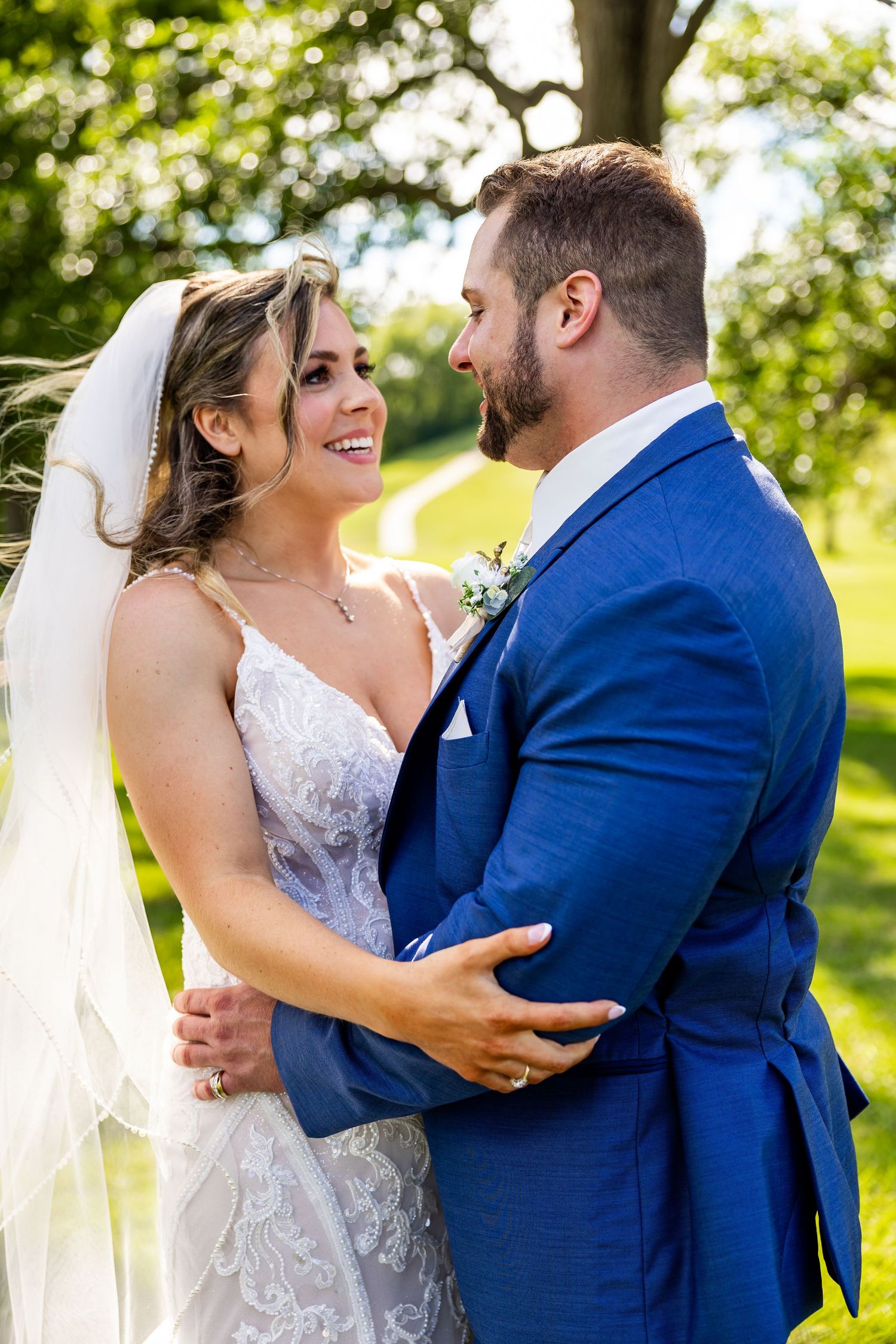 A joyful wedding couple embraces outdoors on a sunny day, with the bride in white and groom in navy blue.