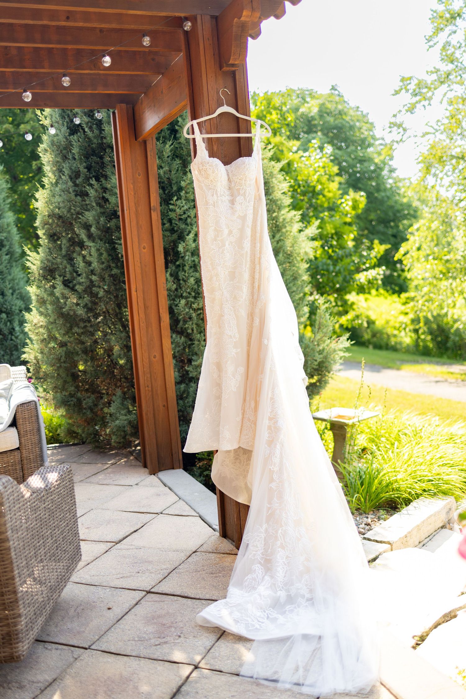 A white lace wedding dress hanging elegantly against a wooden pergola in a garden setting.
