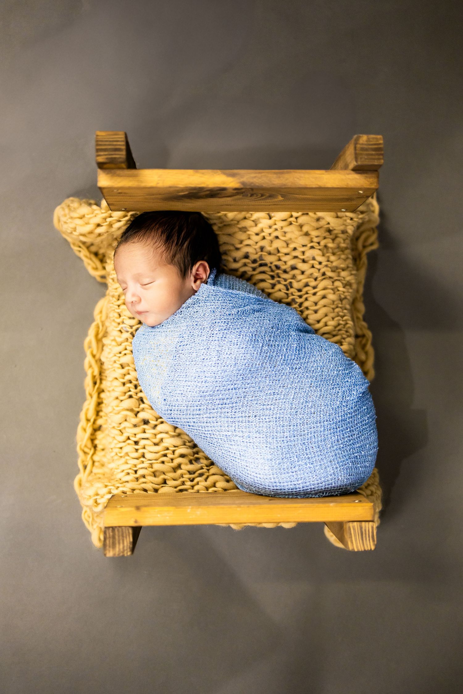 A sleeping baby in a blue blanket resting on a knitted yellow cushion in a wooden basket.