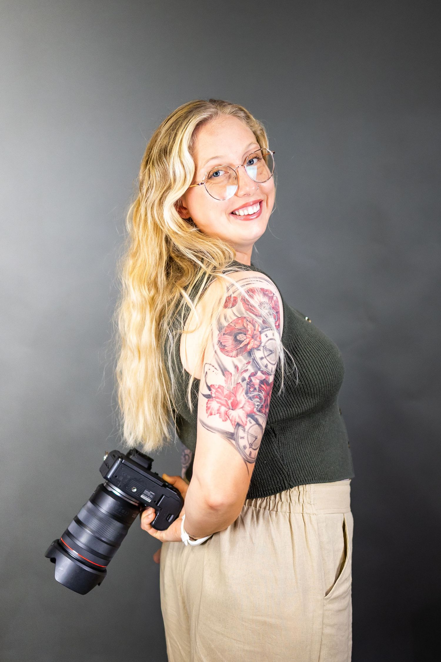 Photographer with colorful arm tattoos poses playfully against grey backdrop while holding a professional camera.