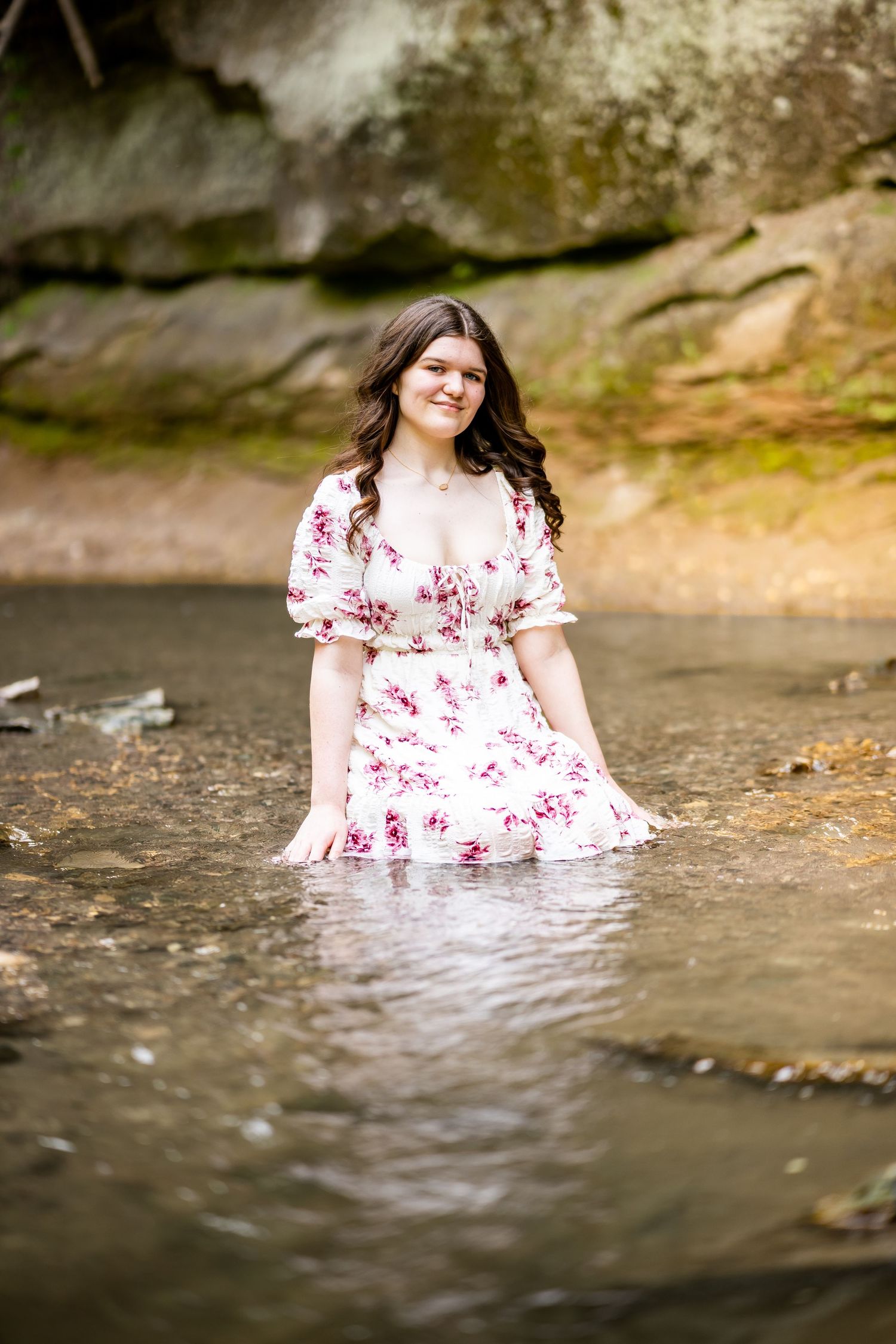 Person in floral dress sitting in shallow creek water for artistic photo sequence.