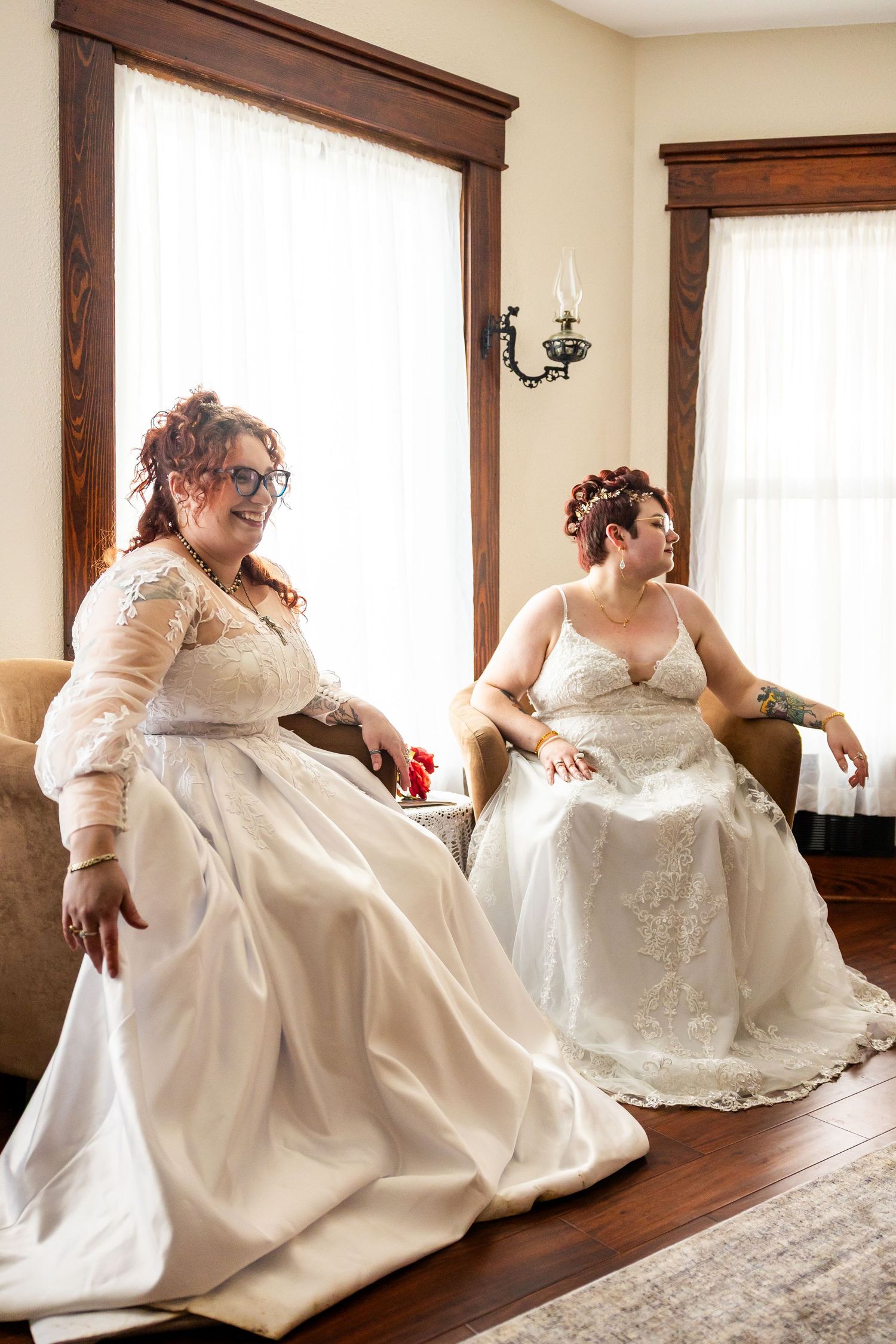 Two brides in white wedding gowns sitting in armchairs by a bright window in an elegant room.