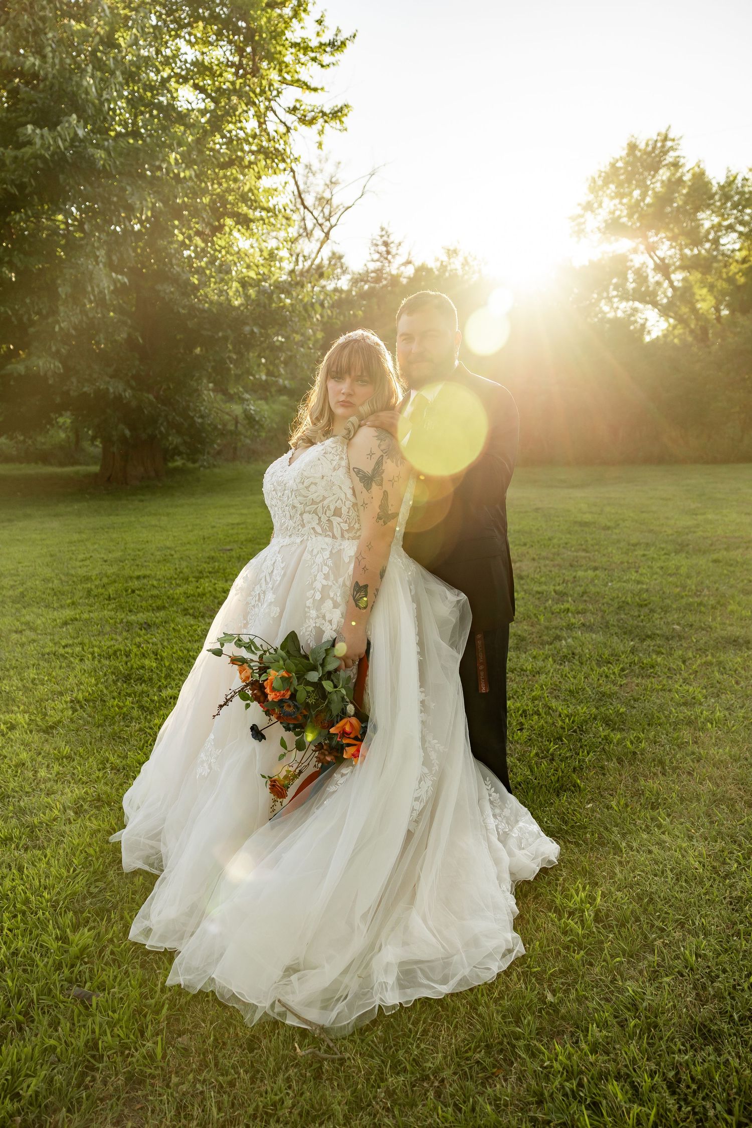Newlywed couple sits together on sunlit grassy lawn during golden hour with autumn wedding bouquet.