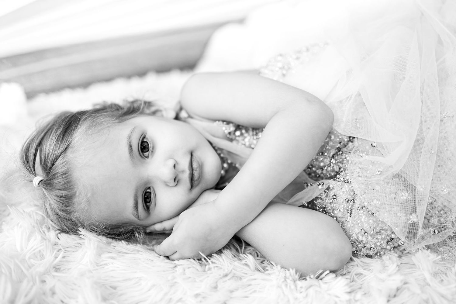 Black and white portrait of a young girl in a sequined dress lying on a fluffy white rug, smiling softly.