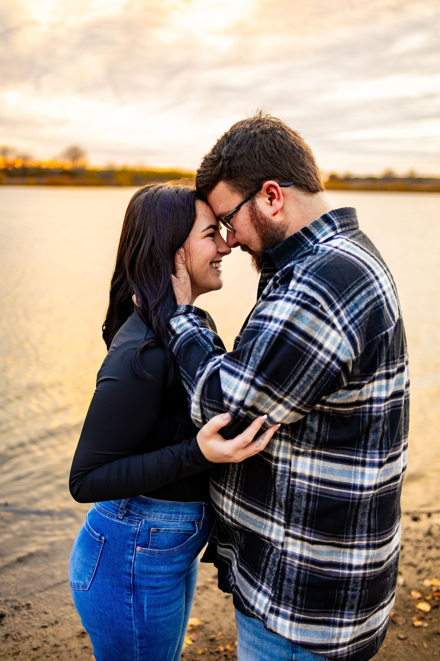 Intimate couple shares tender moment at sunset by a lake wearing casual fall clothing.