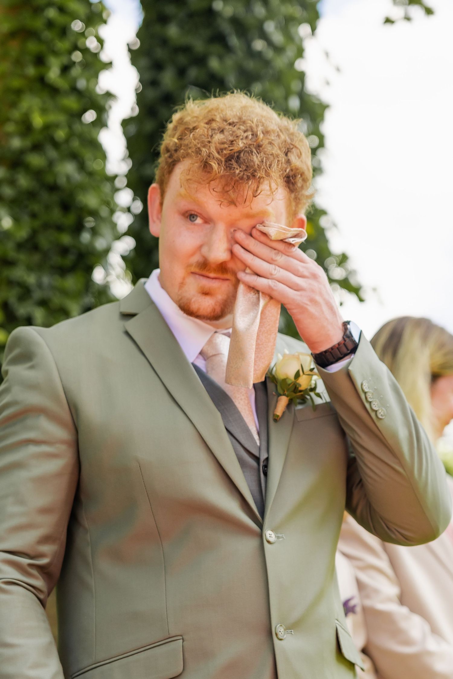 Emotional groom in sage green suit wiping tears at outdoor wedding ceremony with floral boutonniere.