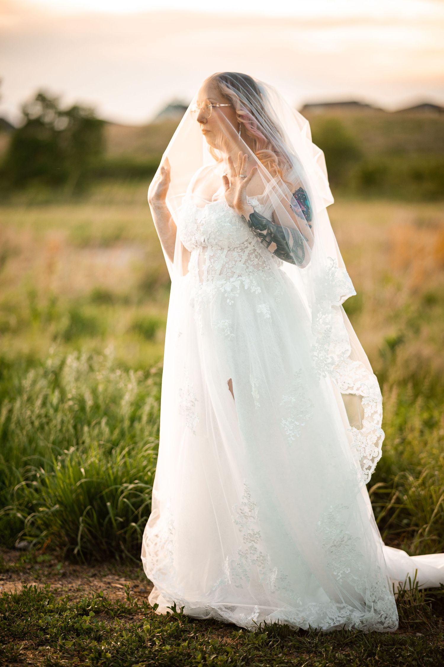 Bride in white lace wedding dress and veil standing in green field at golden sunset.