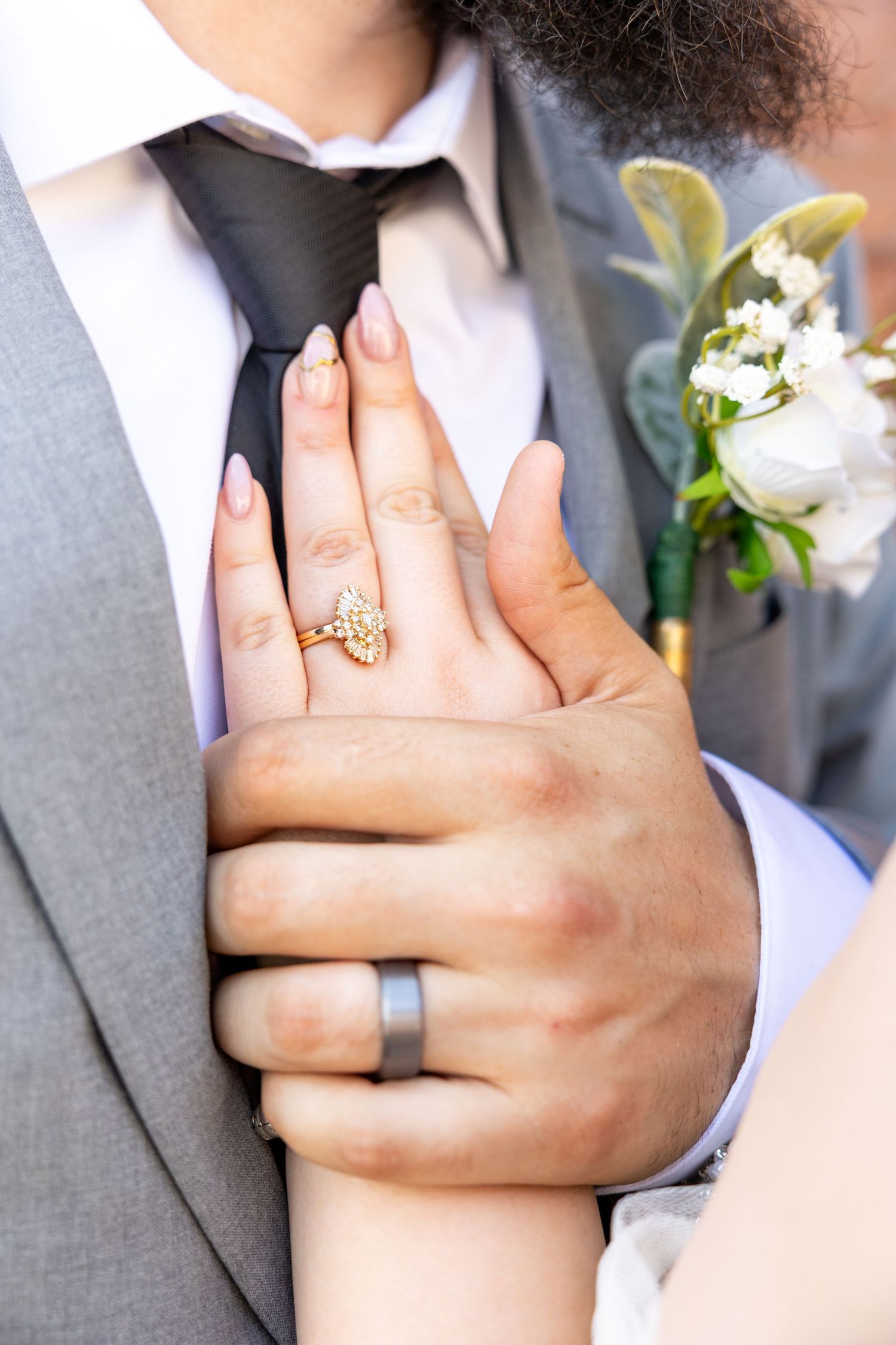 Close-up of hands showing wedding rings with a detailed gold engagement ring and simple silver band against a gray suit.