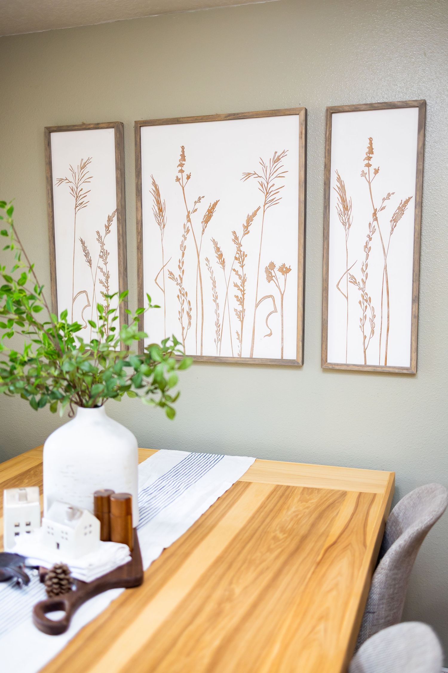 Three framed botanical prints of wheat stalks hang above a wooden table with a white vase and decor items.