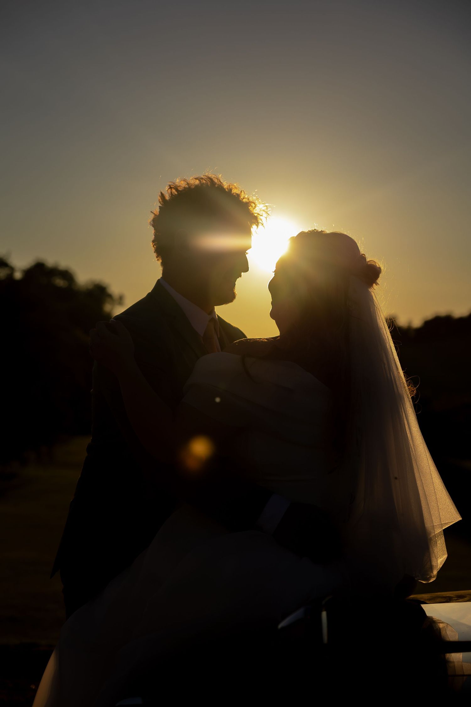 Silhouette of a bride and groom facing each other at sunset, with golden backlight and lens flare creating a romantic scene.