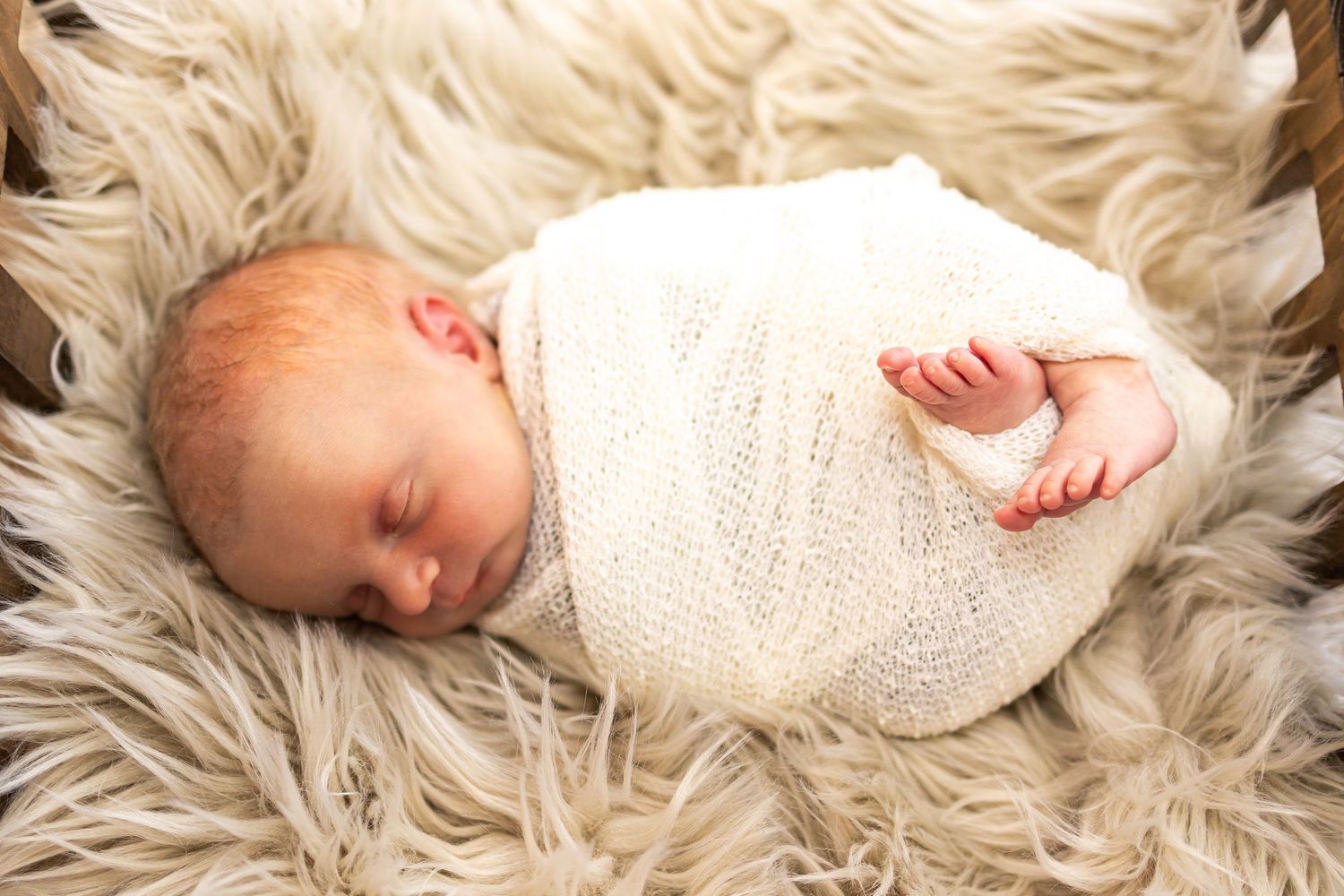 Newborn baby swaddled in white knit wrap sleeping peacefully on a fluffy cream fur blanket.