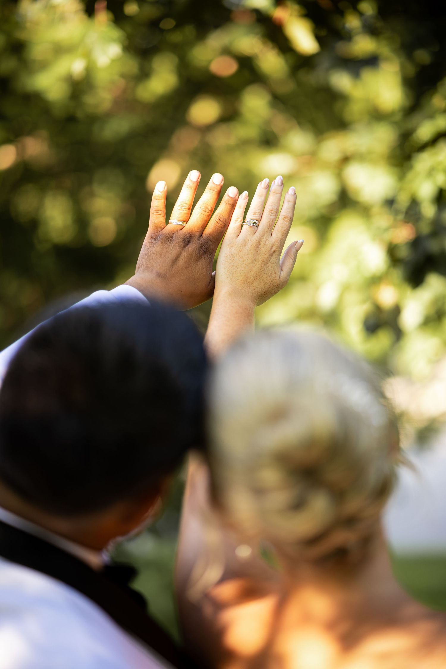 Newlywed couple raising their hands to show wedding rings outdoors with green trees in the background.