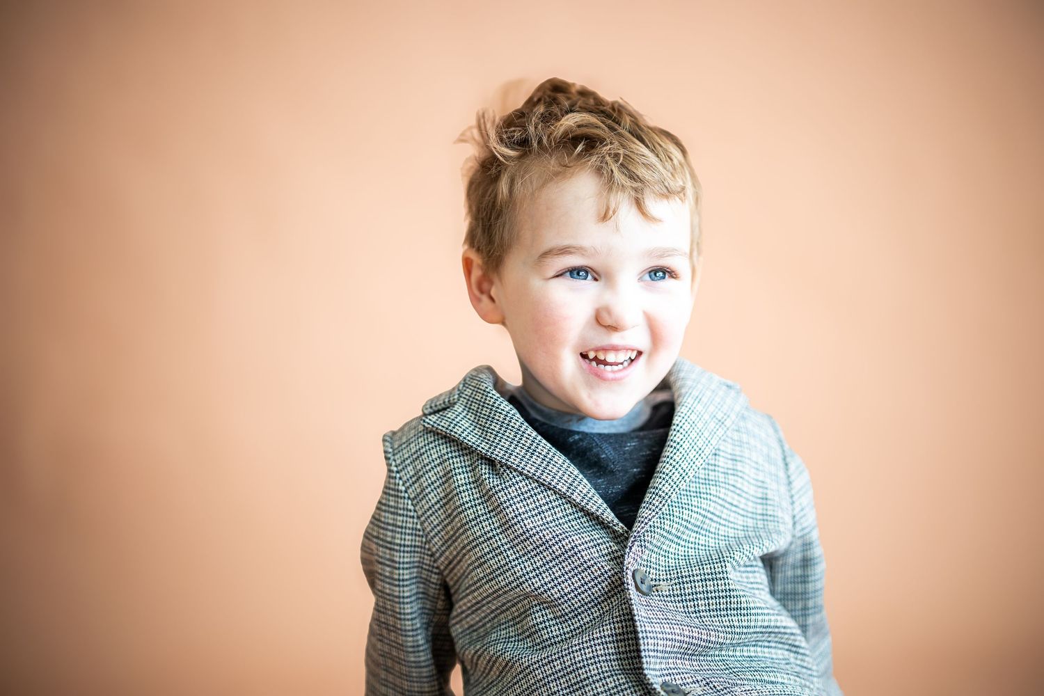 Smiling young boy with curly hair wearing a stylish plaid blazer, posing against a warm beige background.
