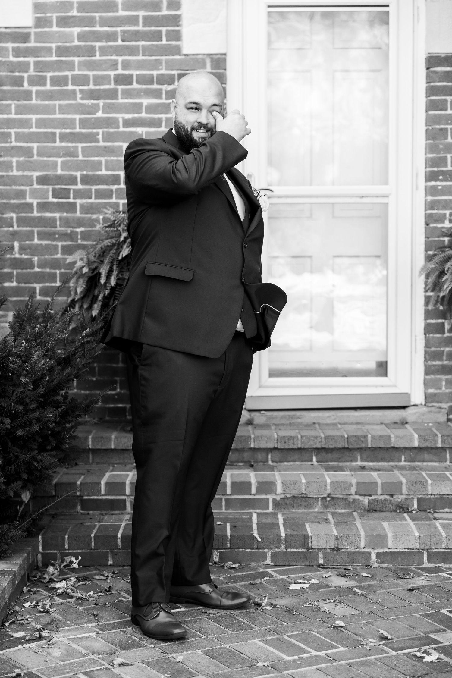 Smiling bald man in black suit poses against brick wall near window in black and white photo.