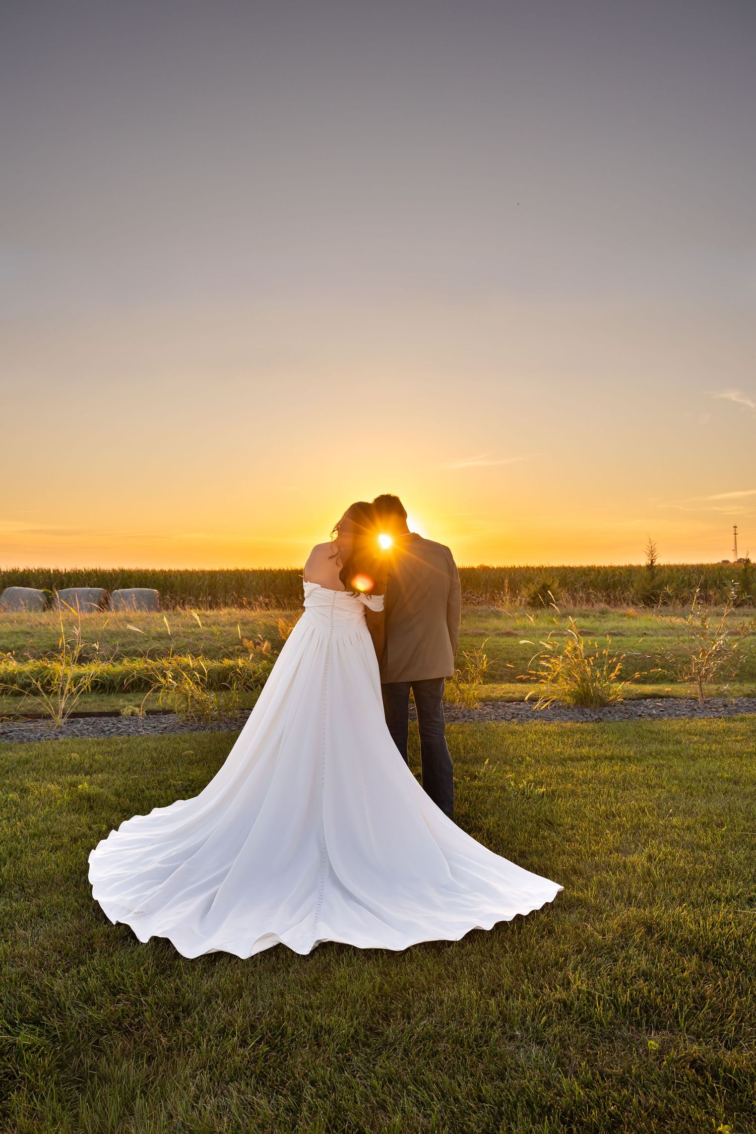 Bride and groom silhouetted against a golden sunset, embracing on a green lawn with a flowing white wedding gown.