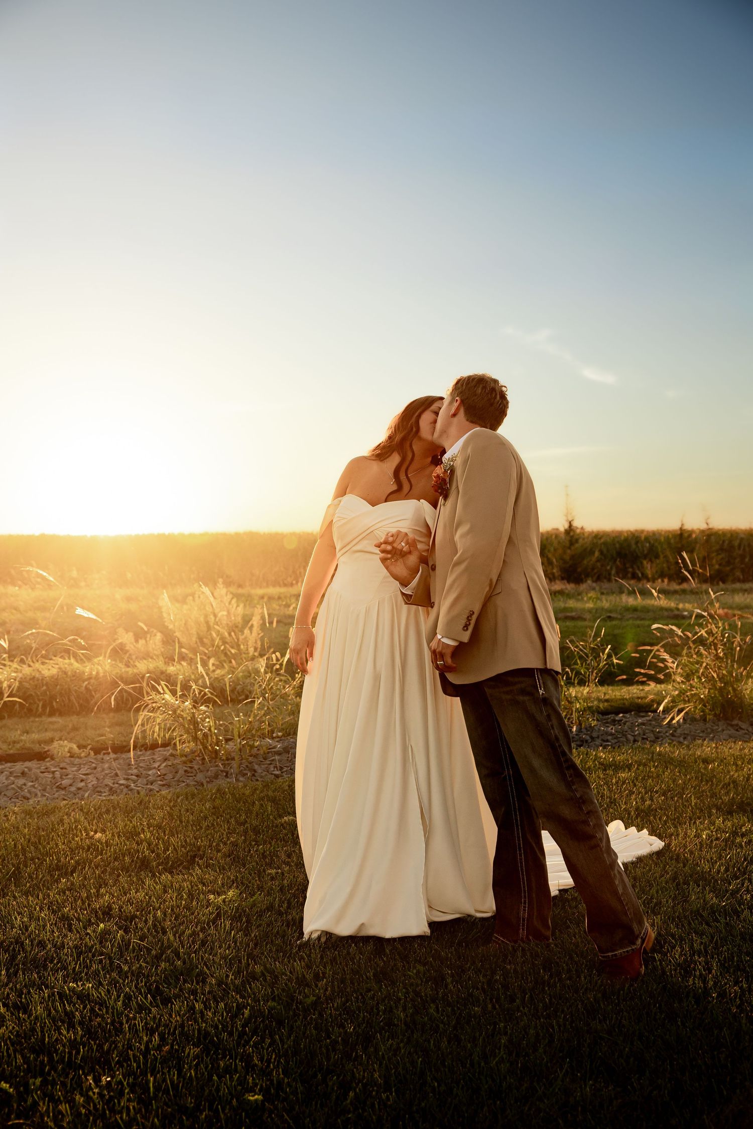A bride and groom share a romantic sunset kiss outdoors, with golden light illuminating their wedding attire.