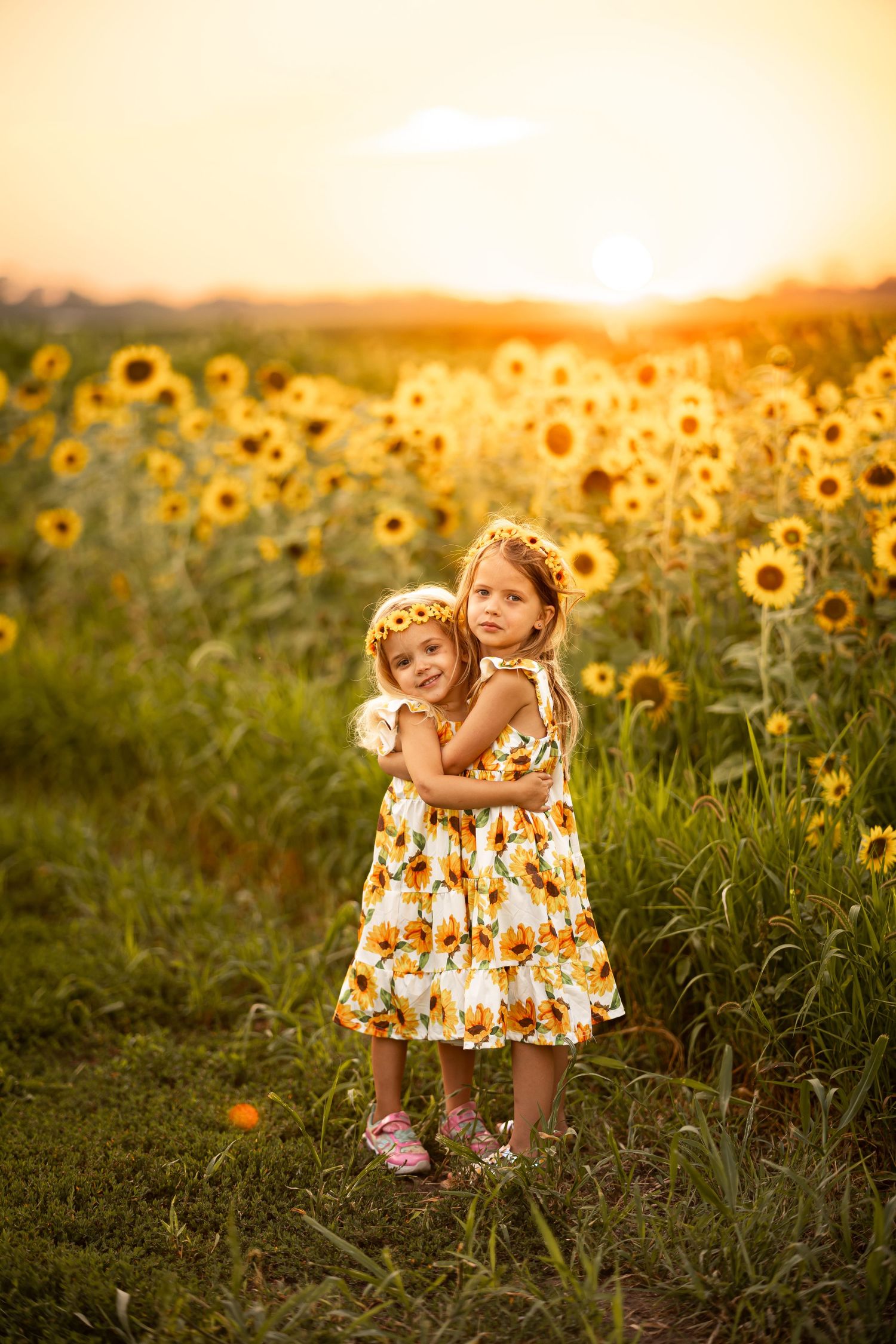 Two young girls in sunflower dresses embrace in a golden sunflower field at sunset, wearing floral wreaths.