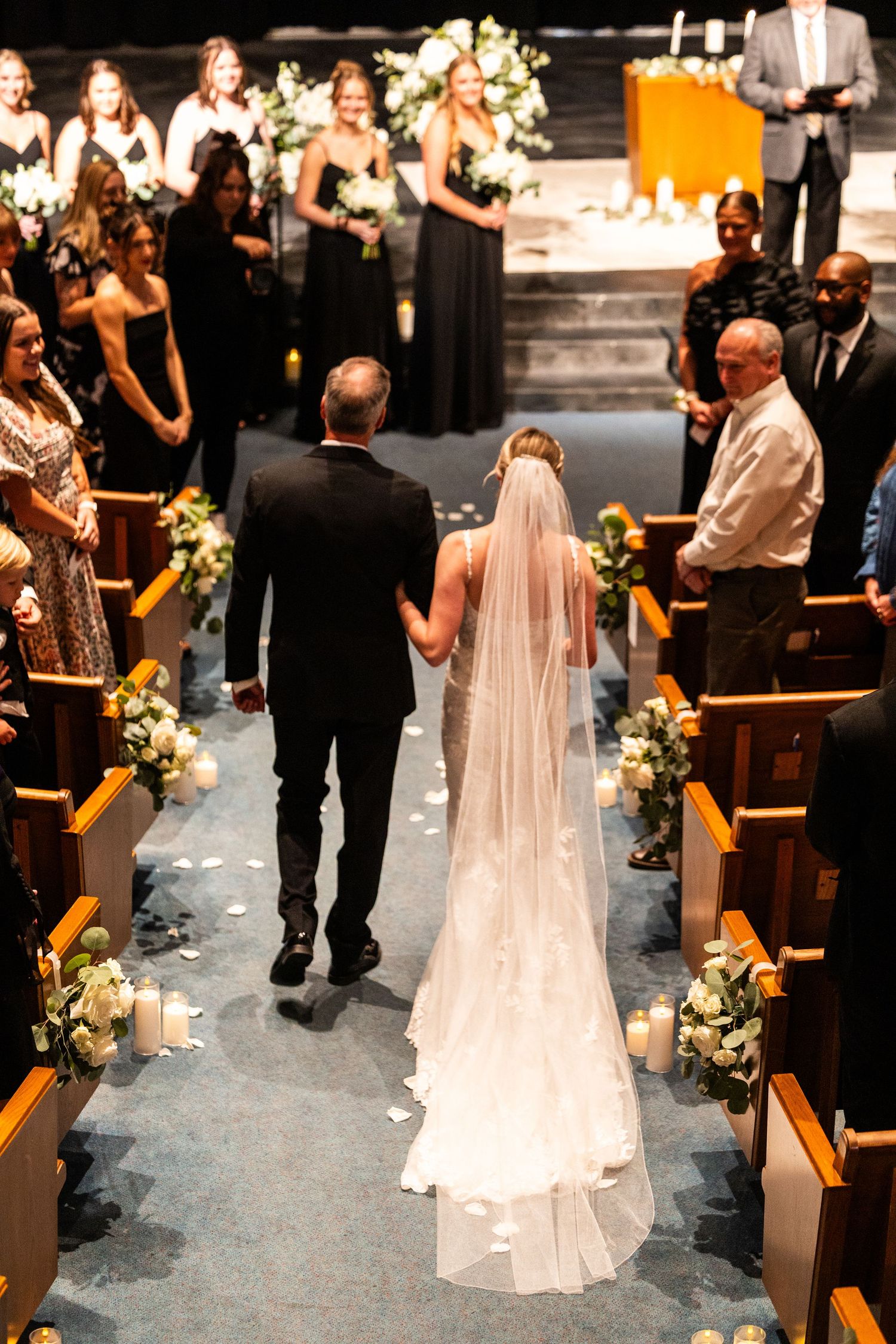 A bride in a flowing veil walks down the church aisle decorated with flowers and candles.