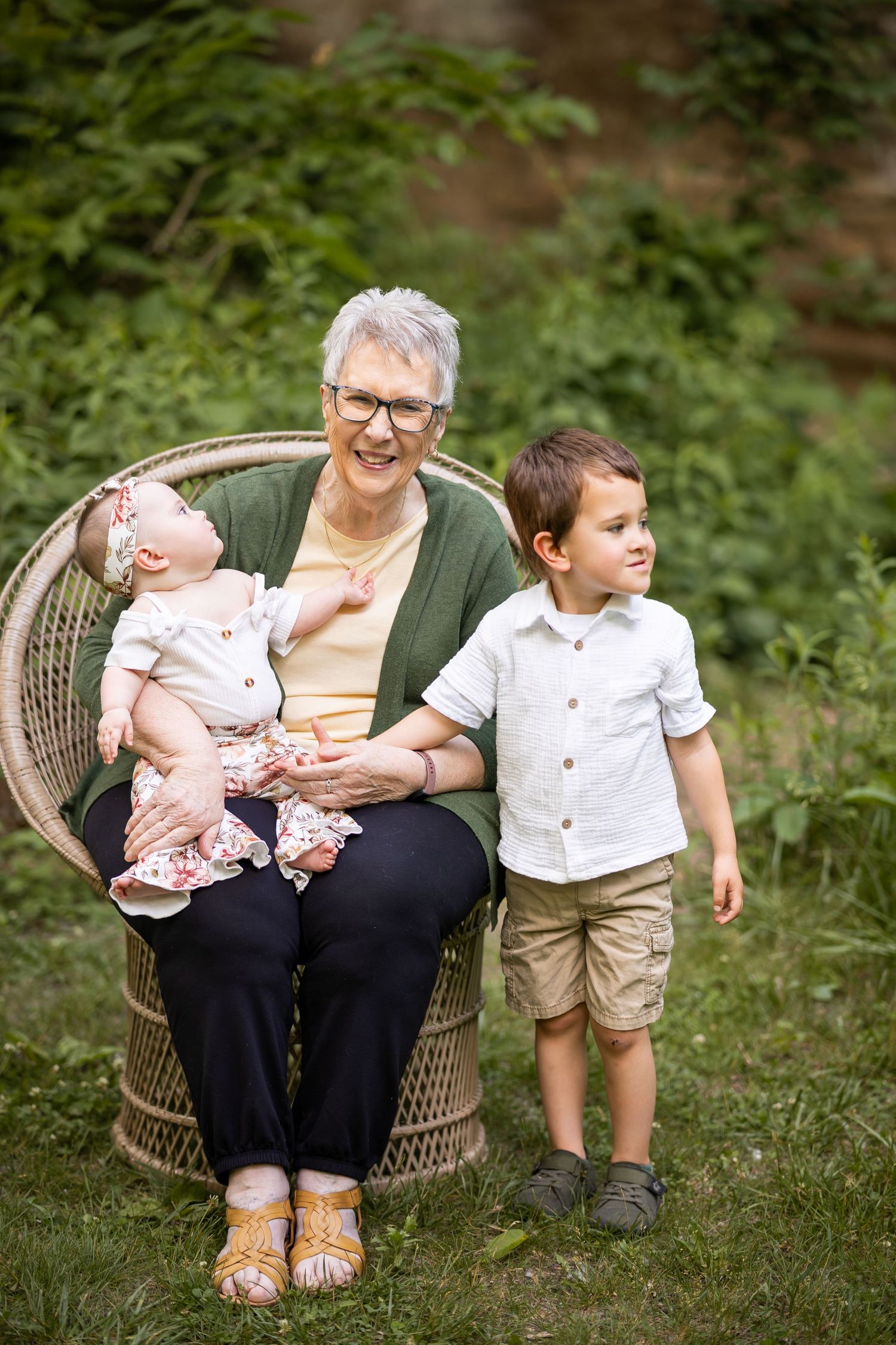 Grandmother sitting in wicker chair outdoors, holding baby girl while young grandson stands beside her smiling.