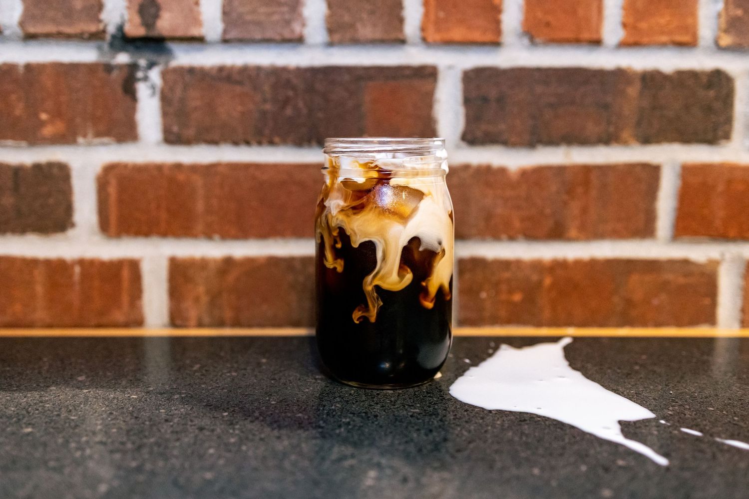 Mason jar filled with iced coffee drink on dark countertop against brick wall.