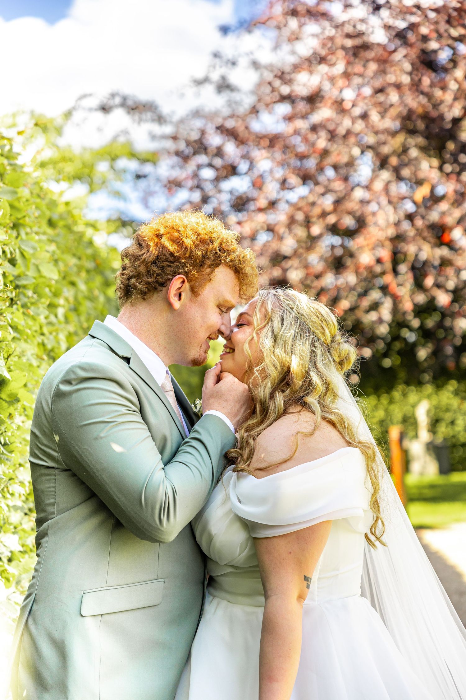 A bride and groom share a romantic kiss outdoors, surrounded by lush green and burgundy trees on their wedding day.