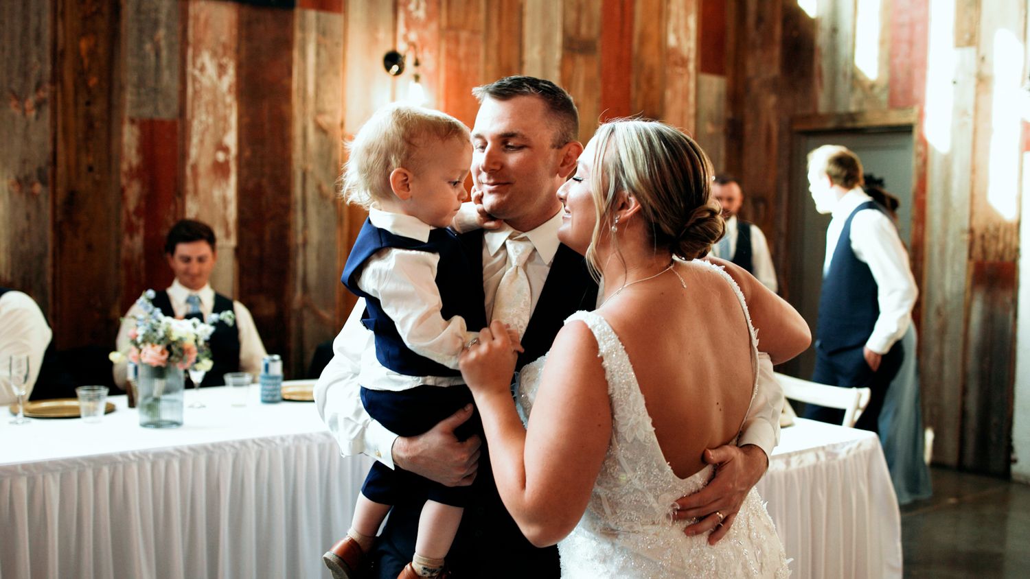 Bride and groom sharing first dance while holding their baby boy in a rustic wedding reception venue.