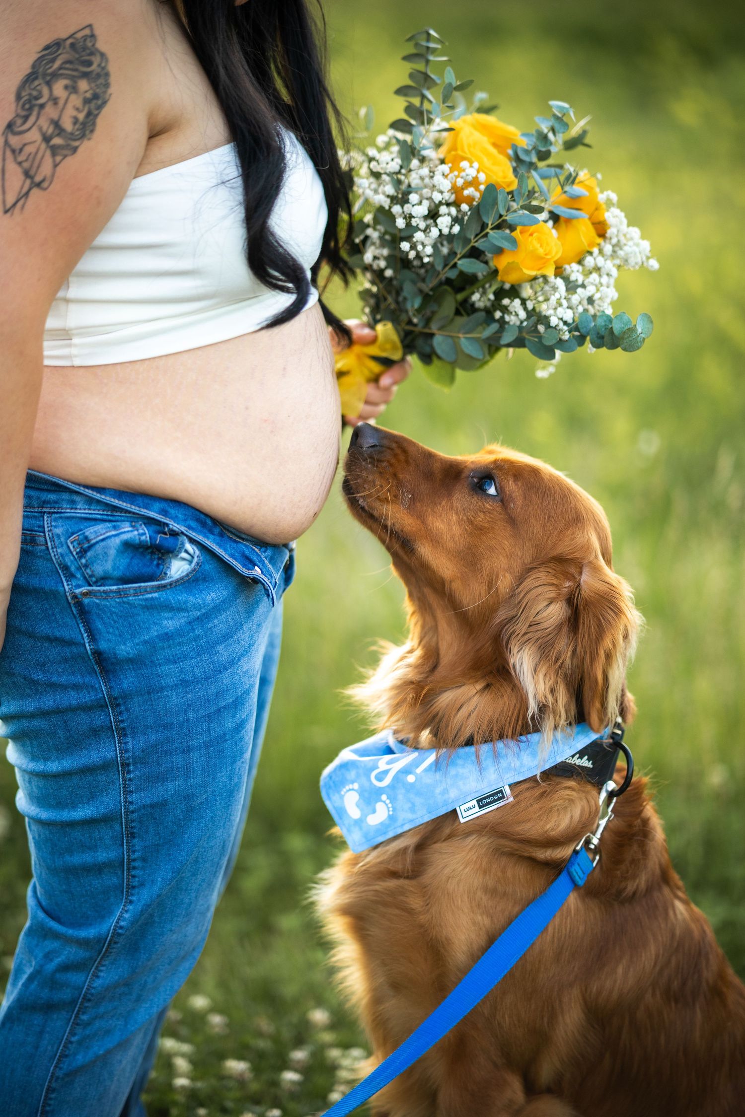 Pregnant woman holding yellow flower bouquet while golden Cocker Spaniel puppy sniffs flowers outdoors.
