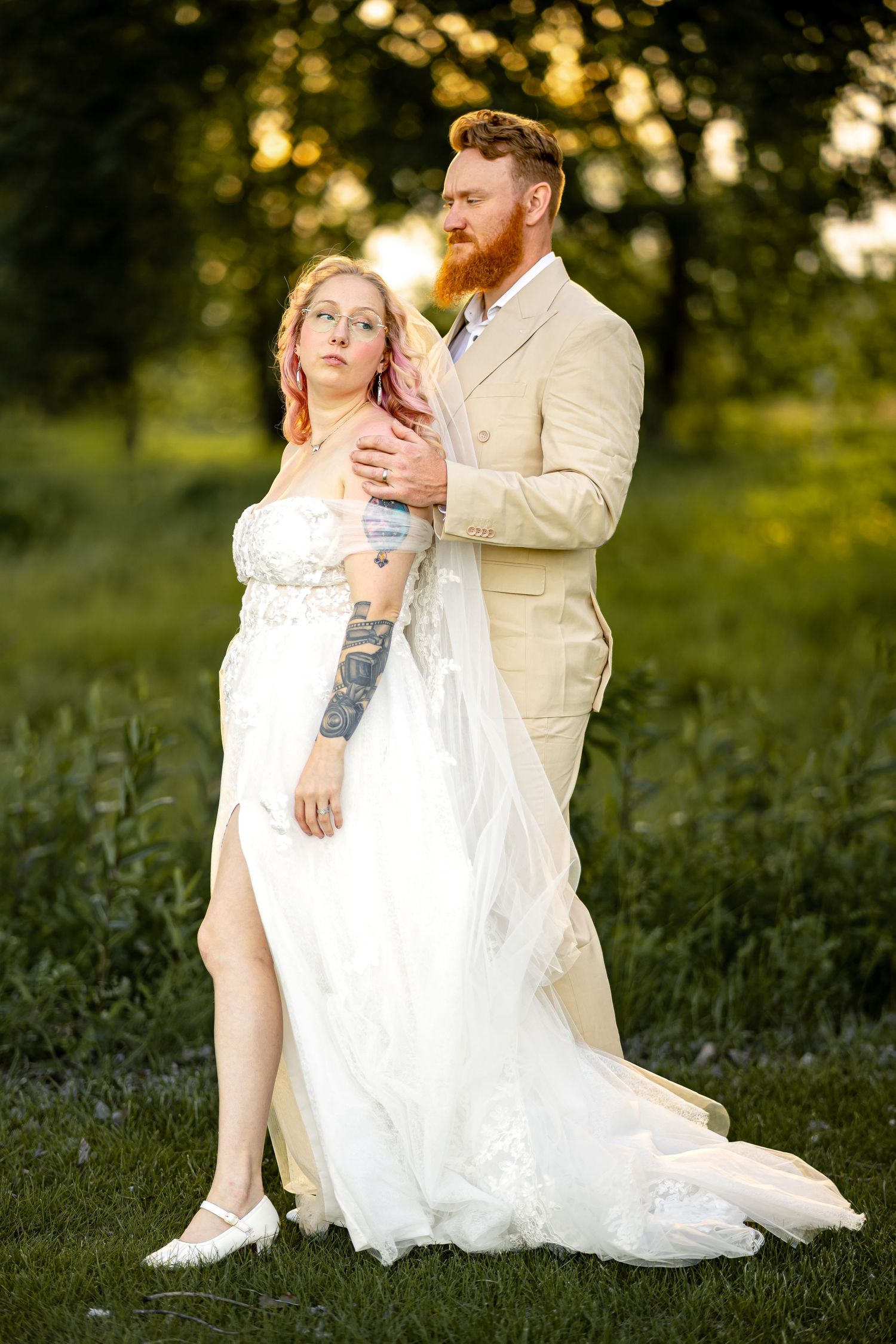 Bride in white lace gown with tattoos stands with bearded groom in tan suit outdoors at golden hour.