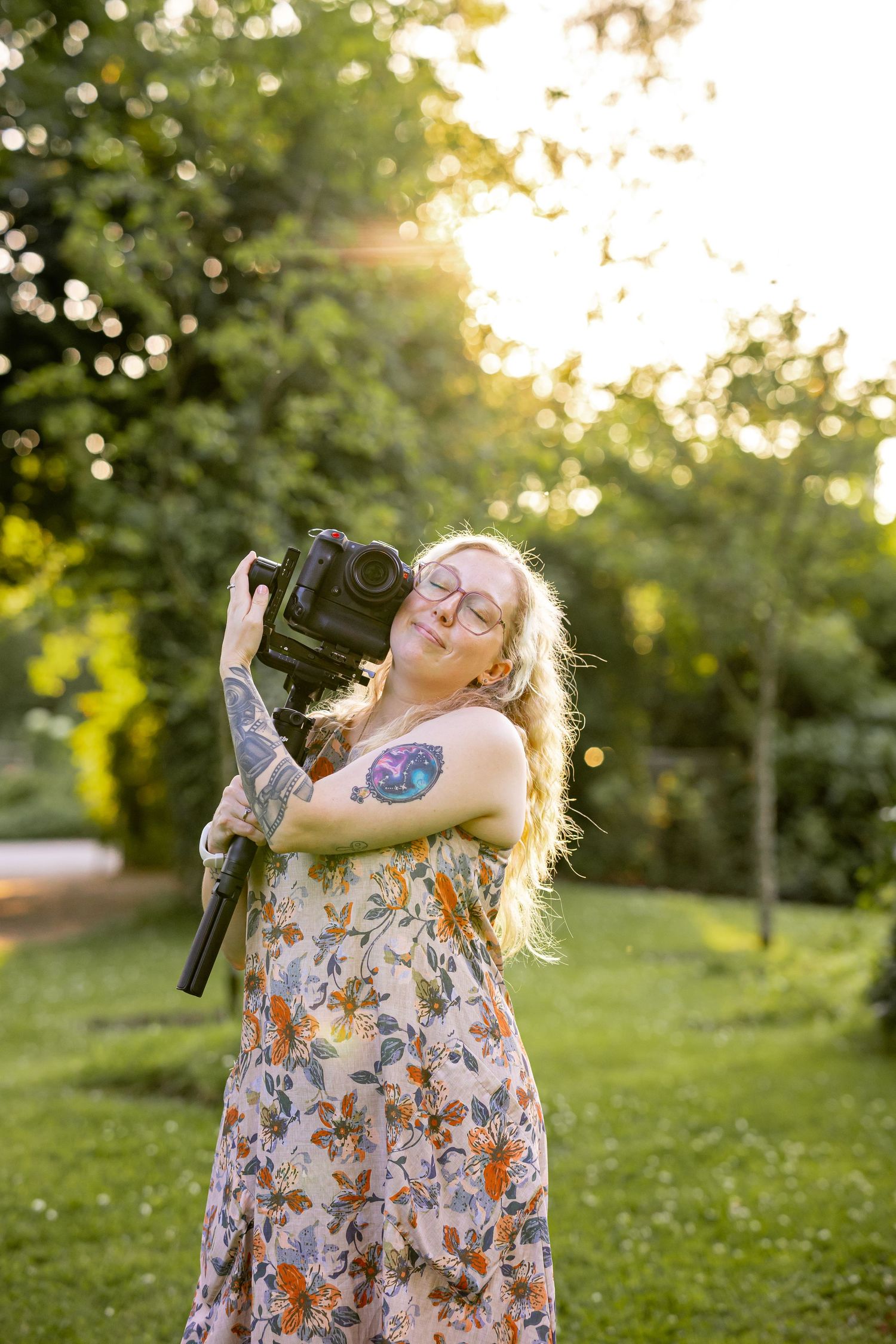 Blonde woman with tattoos in floral dress holding DSLR camera on tripod outdoors in sunny green garden.