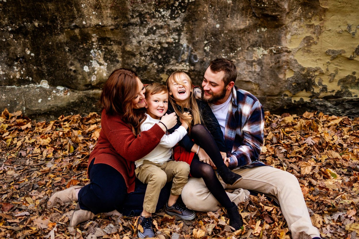 Happy family laughing together while sitting on autumn leaves against stone wall.