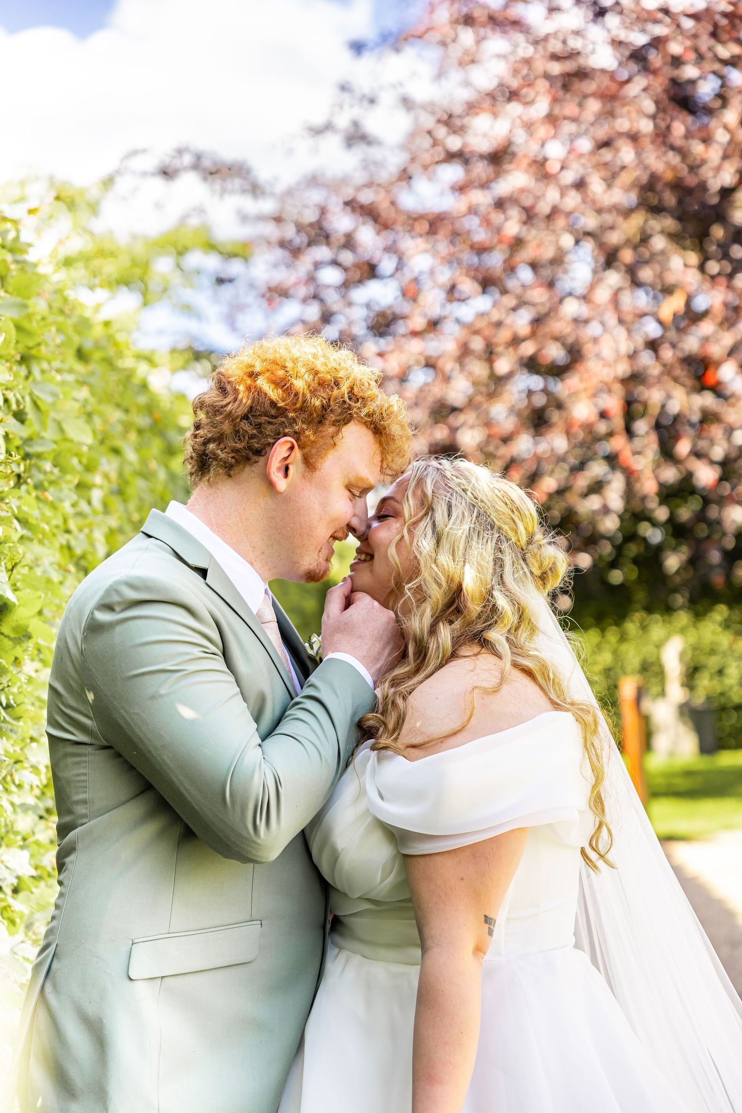 Bride and groom sharing a romantic moment outdoors, groom in sage suit with curly red hair, bride in white off-shoulder gown.