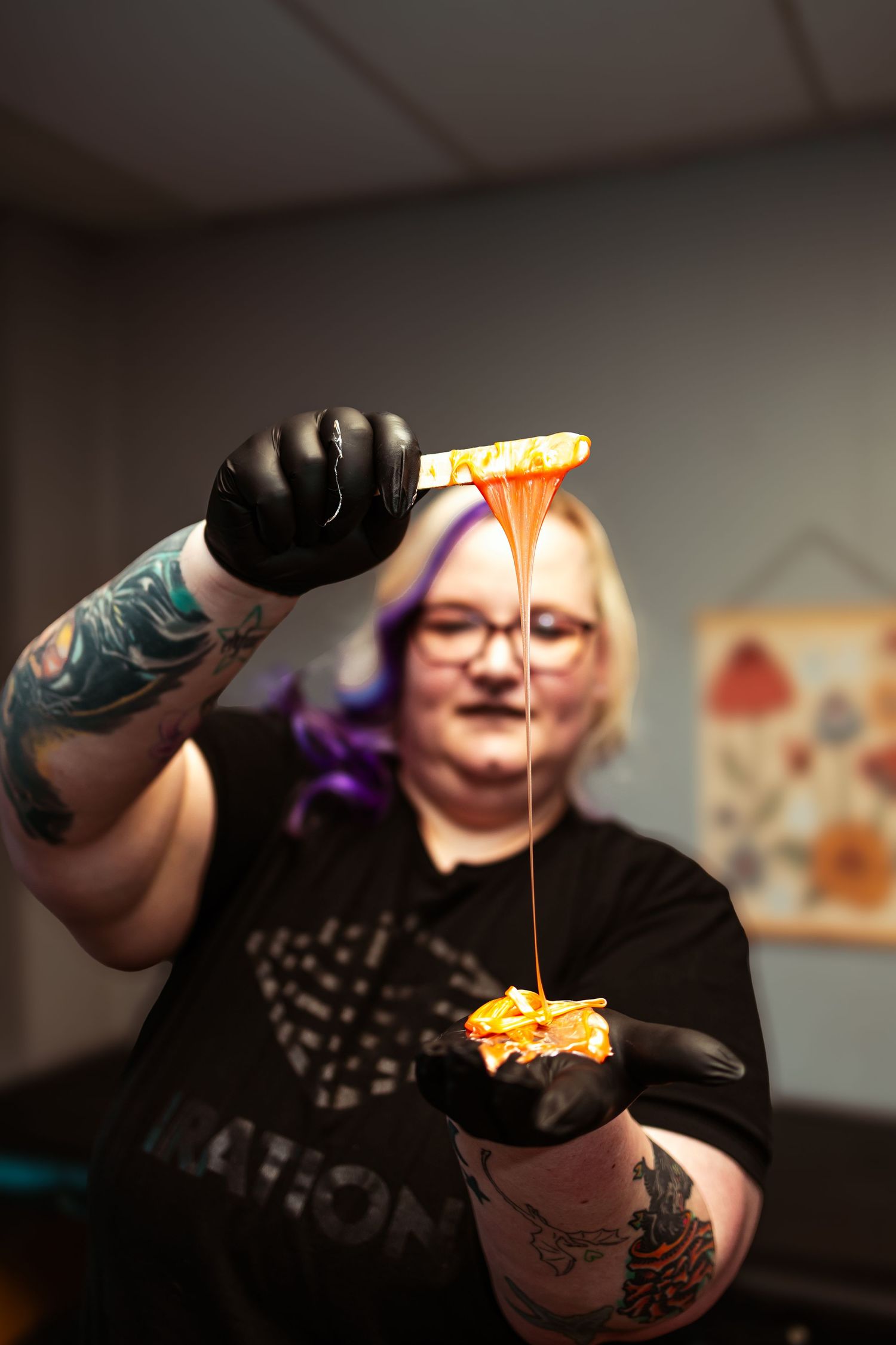 Tattooed woman with purple hair drizzling orange wax from a patterned stick over a golden wax ball in a salon.