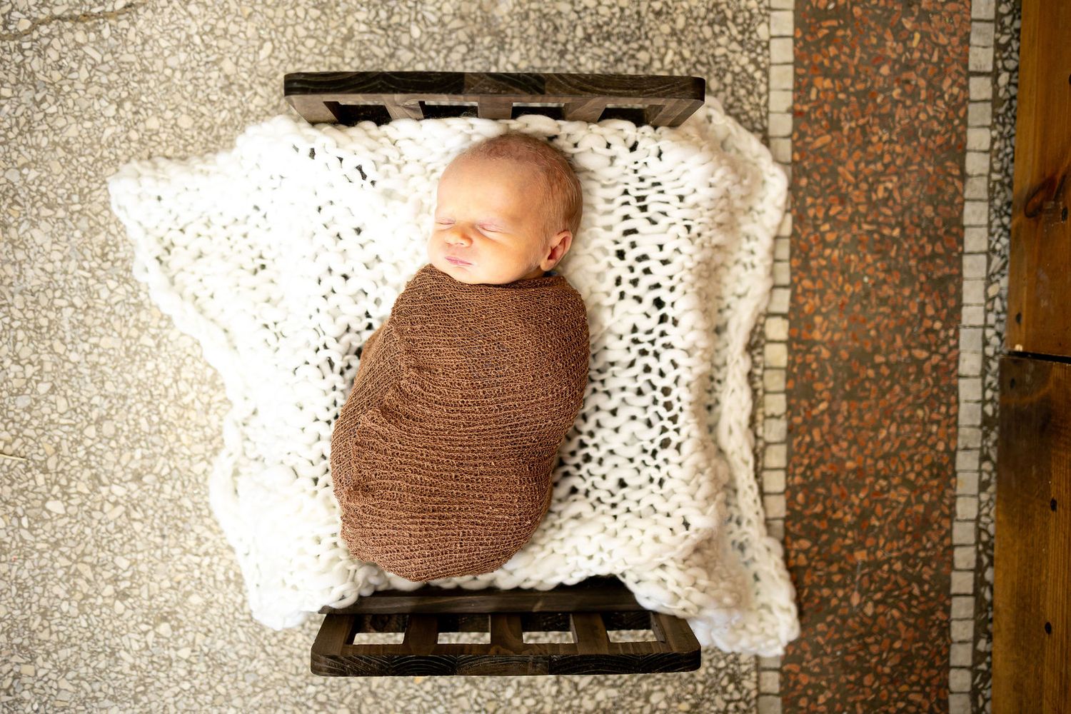 Newborn baby swaddled in brown wrap sleeping on white crochet blanket on rustic wooden pallet bed.