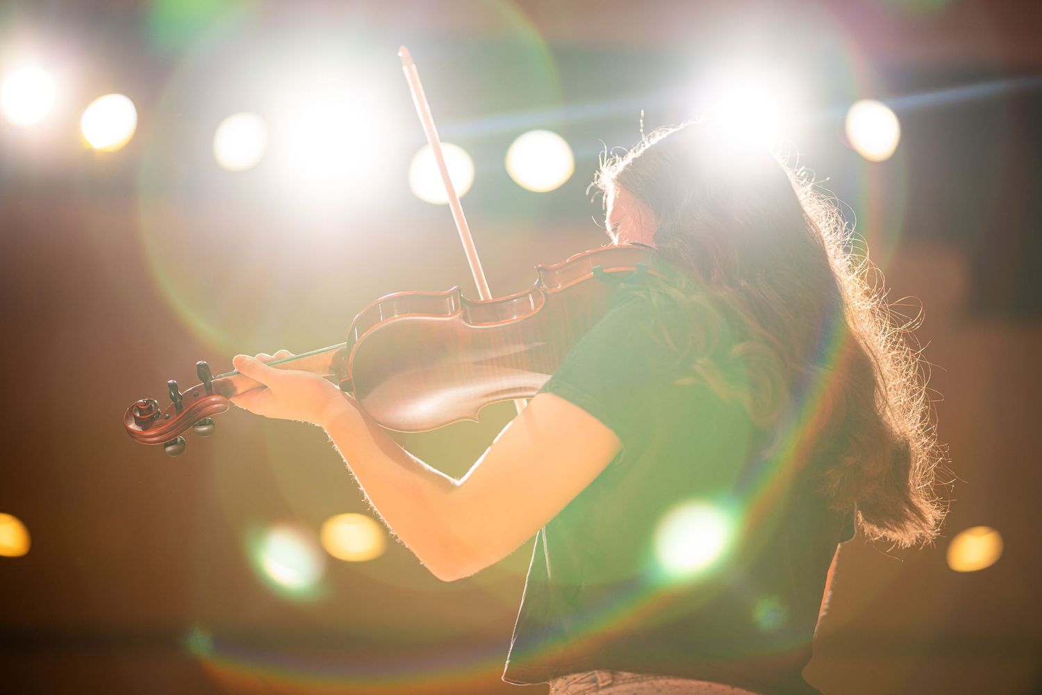 Musician playing violin on stage under bright spotlights with lens flare effect during live performance.