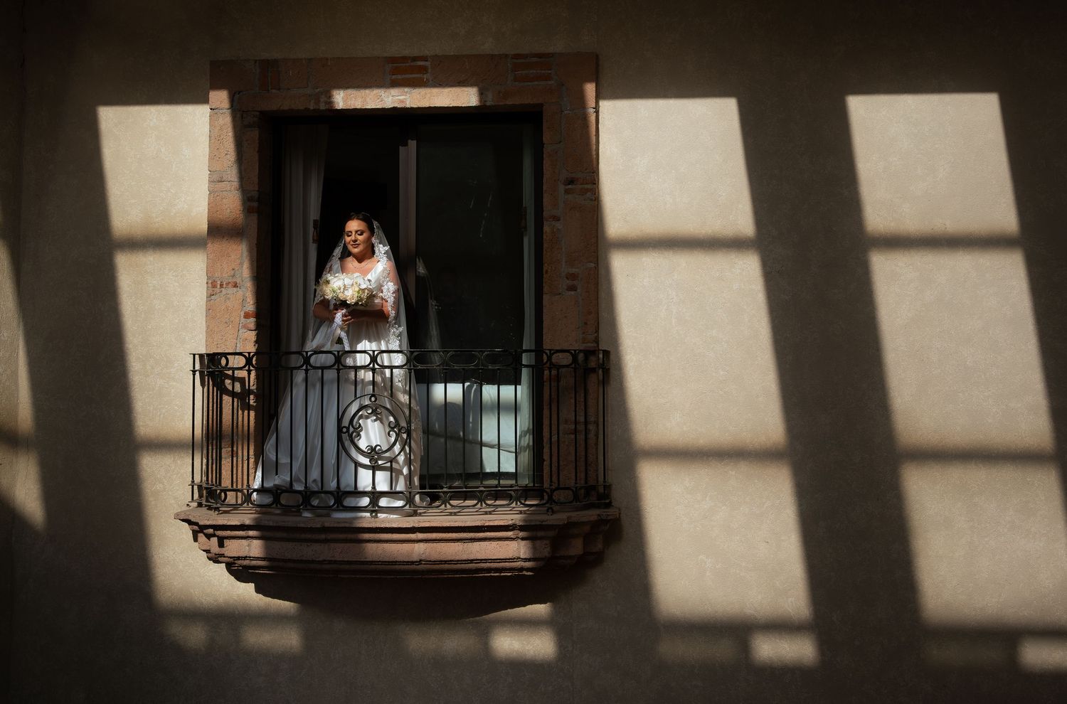 Bride in wedding dress standing on a sunlit balcony, holding bouquet, with dramatic shadows on building facade.