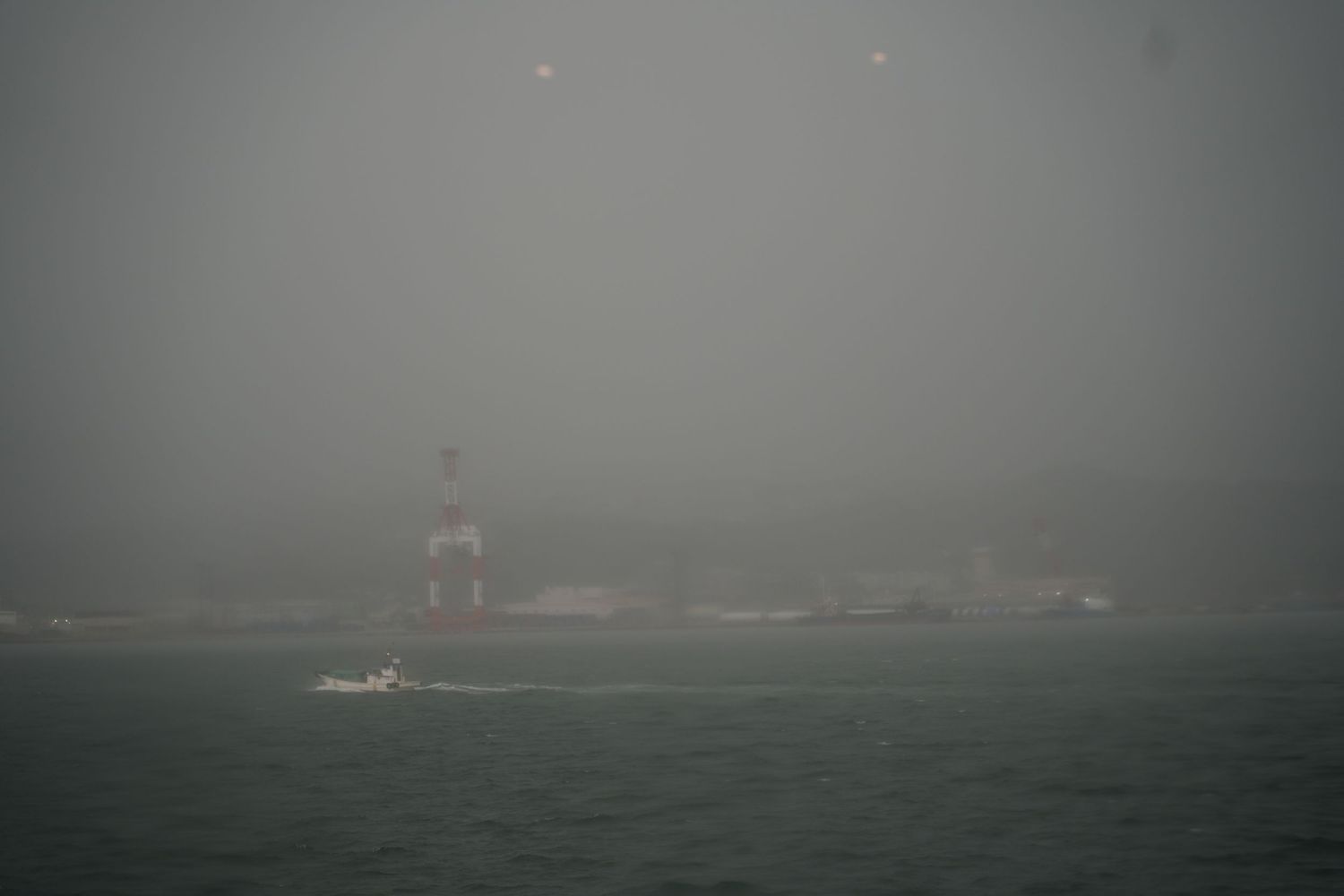 Foggy harbor scene with a red lighthouse and small boat navigating misty waters on an overcast day.