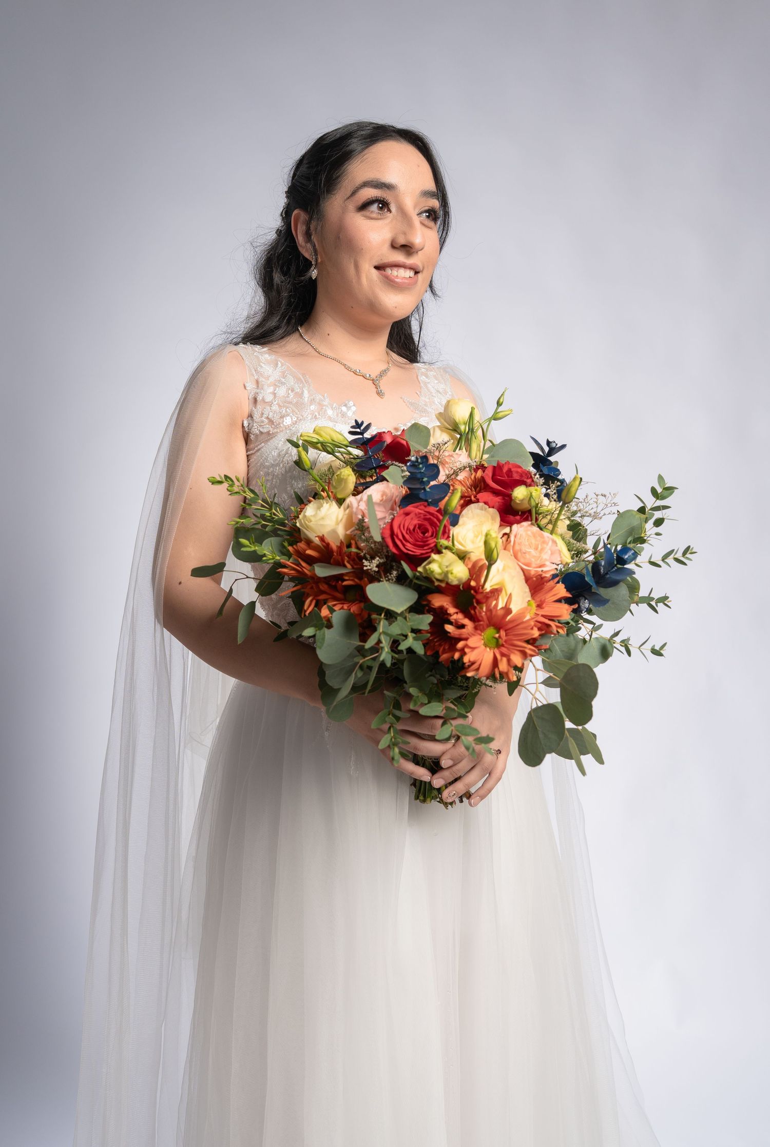Bride in white lace gown holding colorful bouquet of orange, red, and blue flowers with eucalyptus greenery.