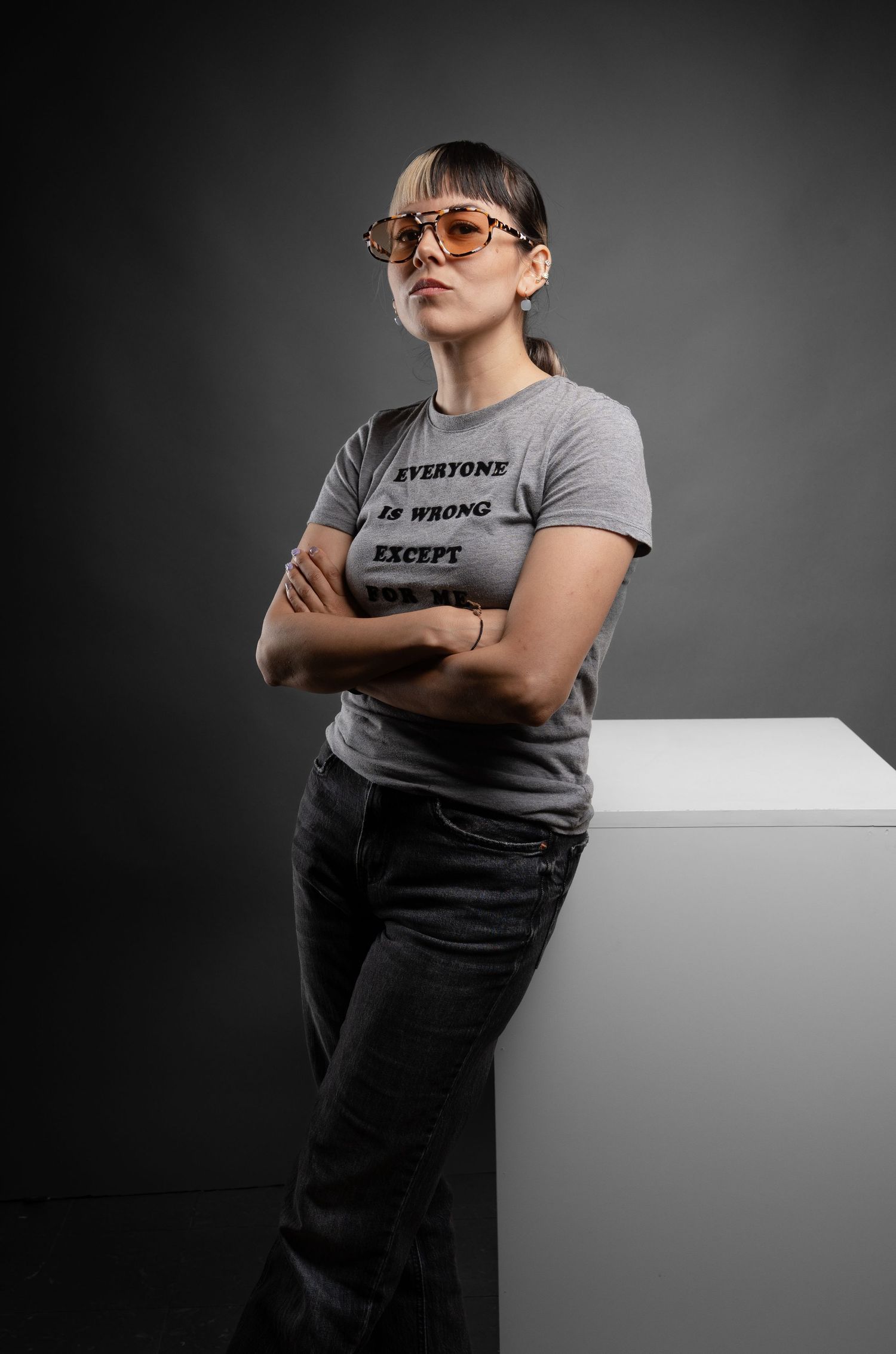 Person in casual attire posing with attitude on white platform against dark background.