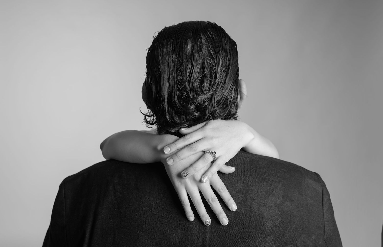 Black and white photo of a person embracing another from behind, showing a ringed hand resting on their shoulder.