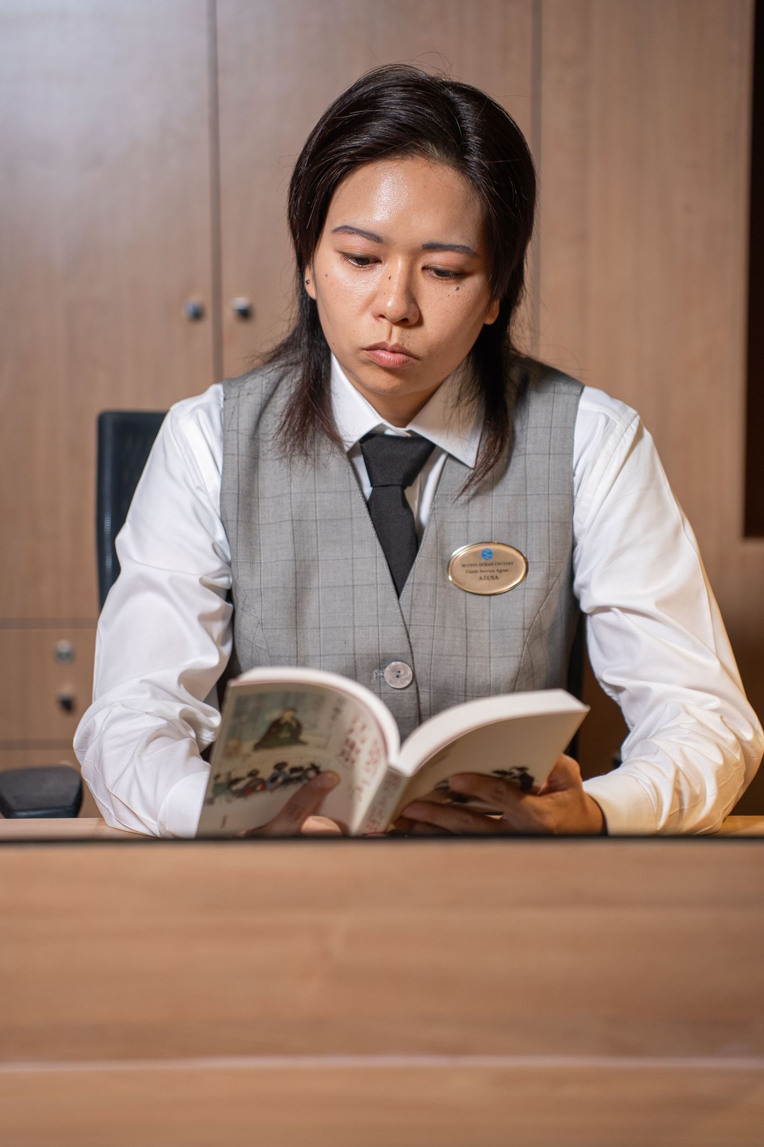 Staff member in grey vest and white shirt reading documents at wooden desk.