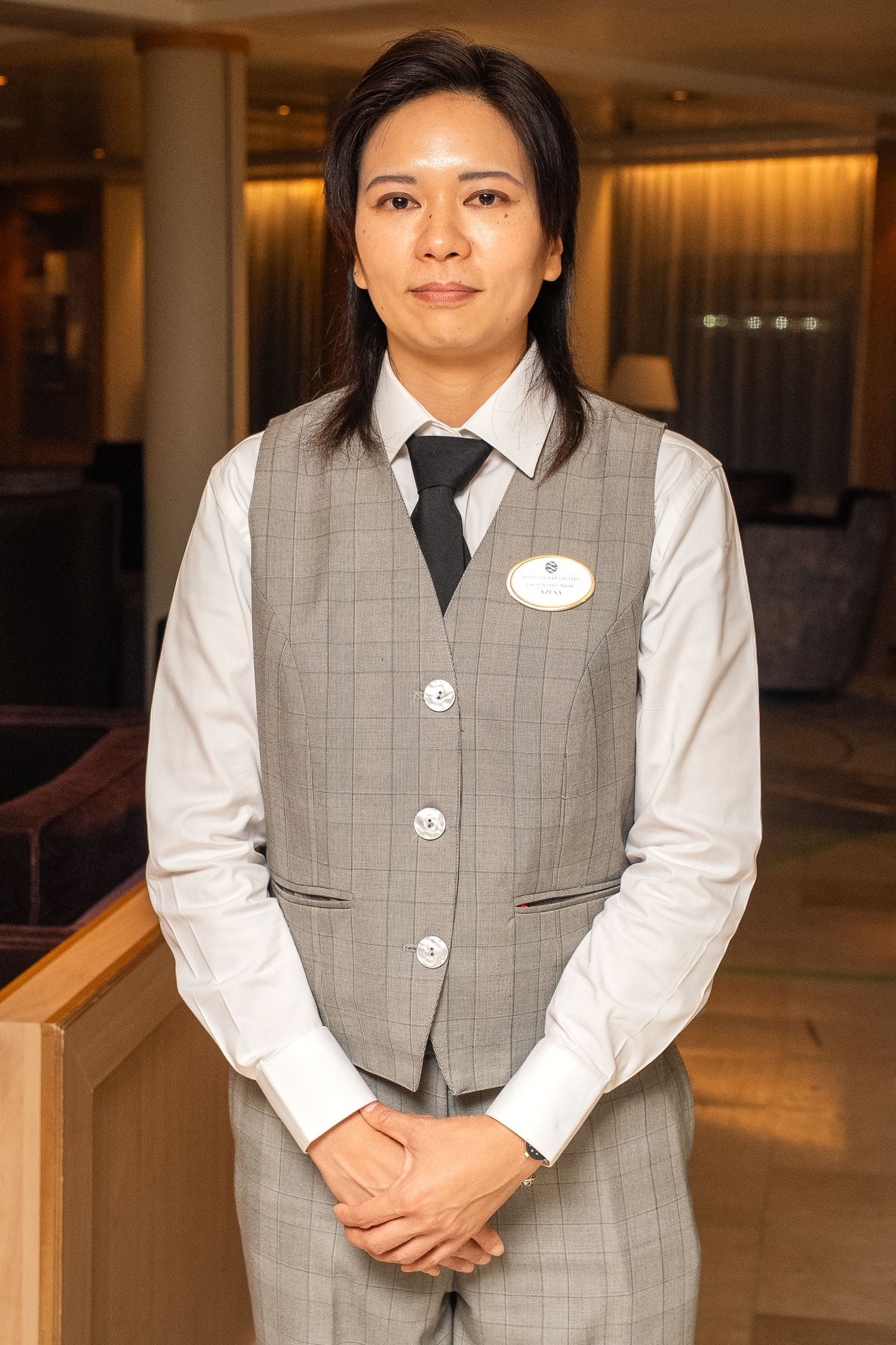 Hotel staff member wearing grey vest and black tie standing in elegant lobby area.